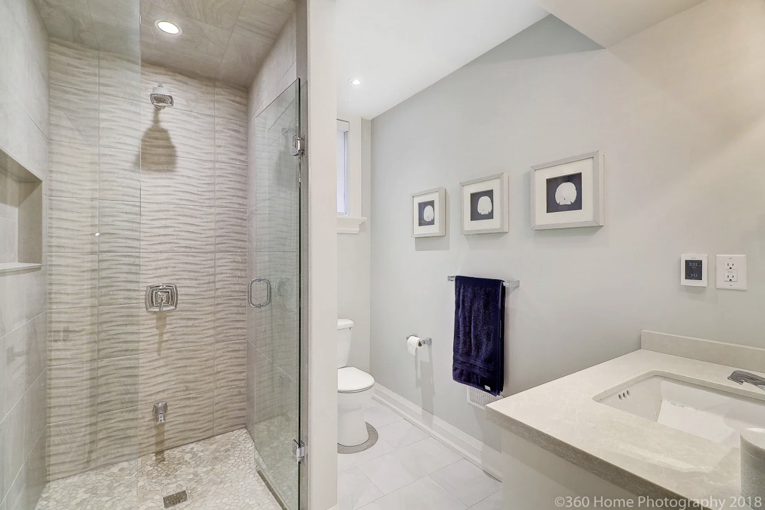 Modern bathroom with a walk-in shower with textured beige tiles, a glass door, and a silver handle. The room has white walls with three framed black and white prints, a purple towel hanging on a silver towel bar, and a white vanity with a sink. There