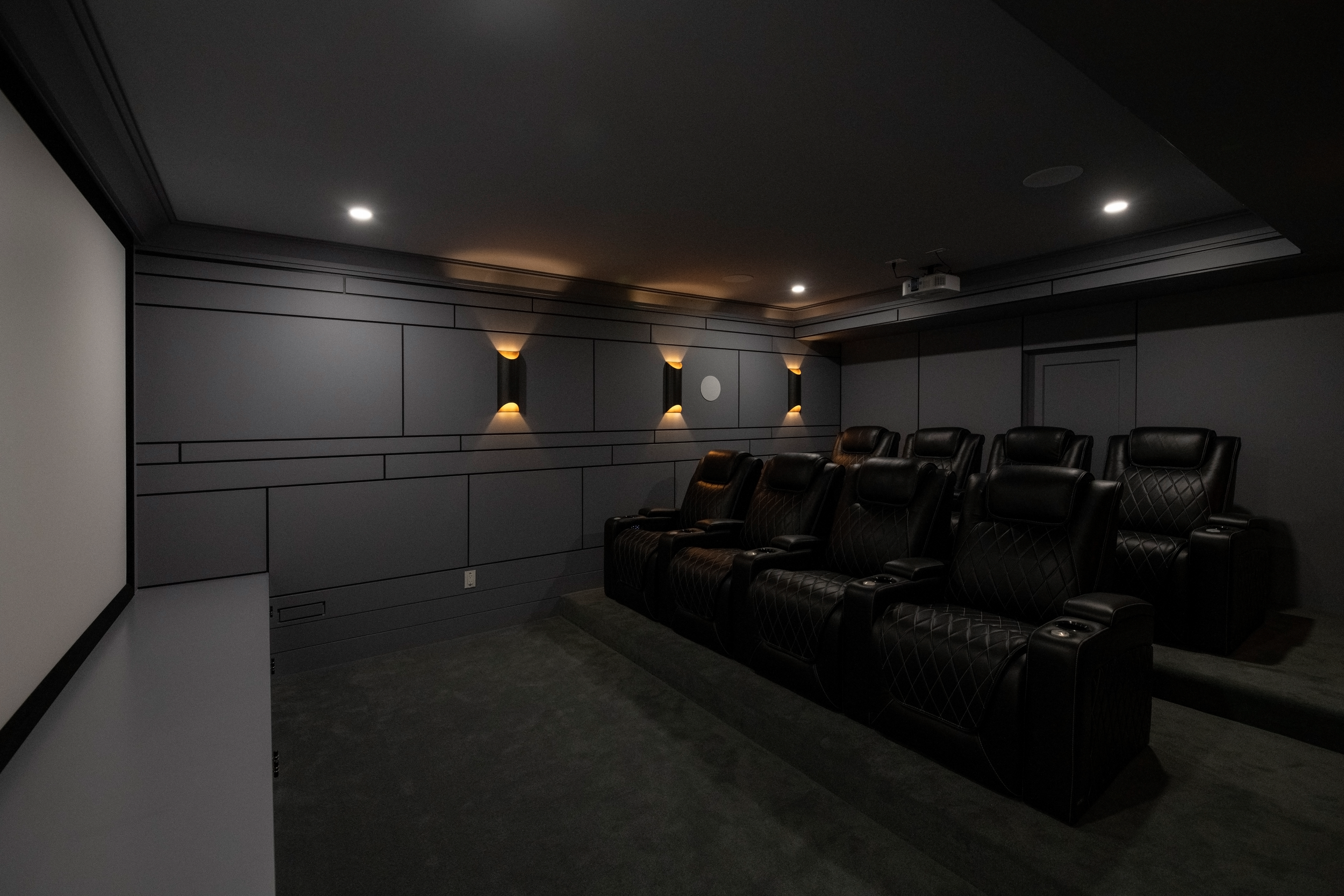 Darkened movie theater room with black leather chairs arranged in rows, wall-mounted lights, and a projector mounted on the ceiling.