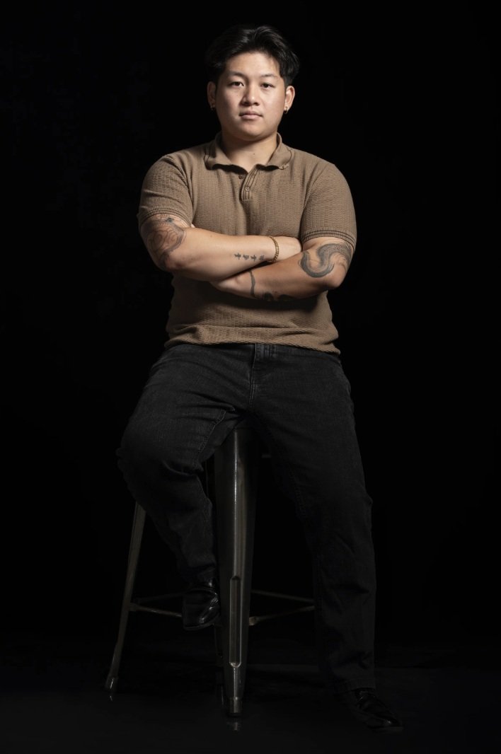 A young Asian man with black hair and tattoos on both arms, sitting on a stool against a black background, wearing a brown polo shirt and black pants, looking directly at the camera with crossed arms.