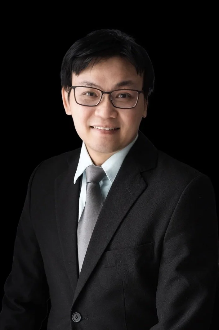 A young man with glasses wearing a black suit, white shirt, and gray tie smiling against a black background.
