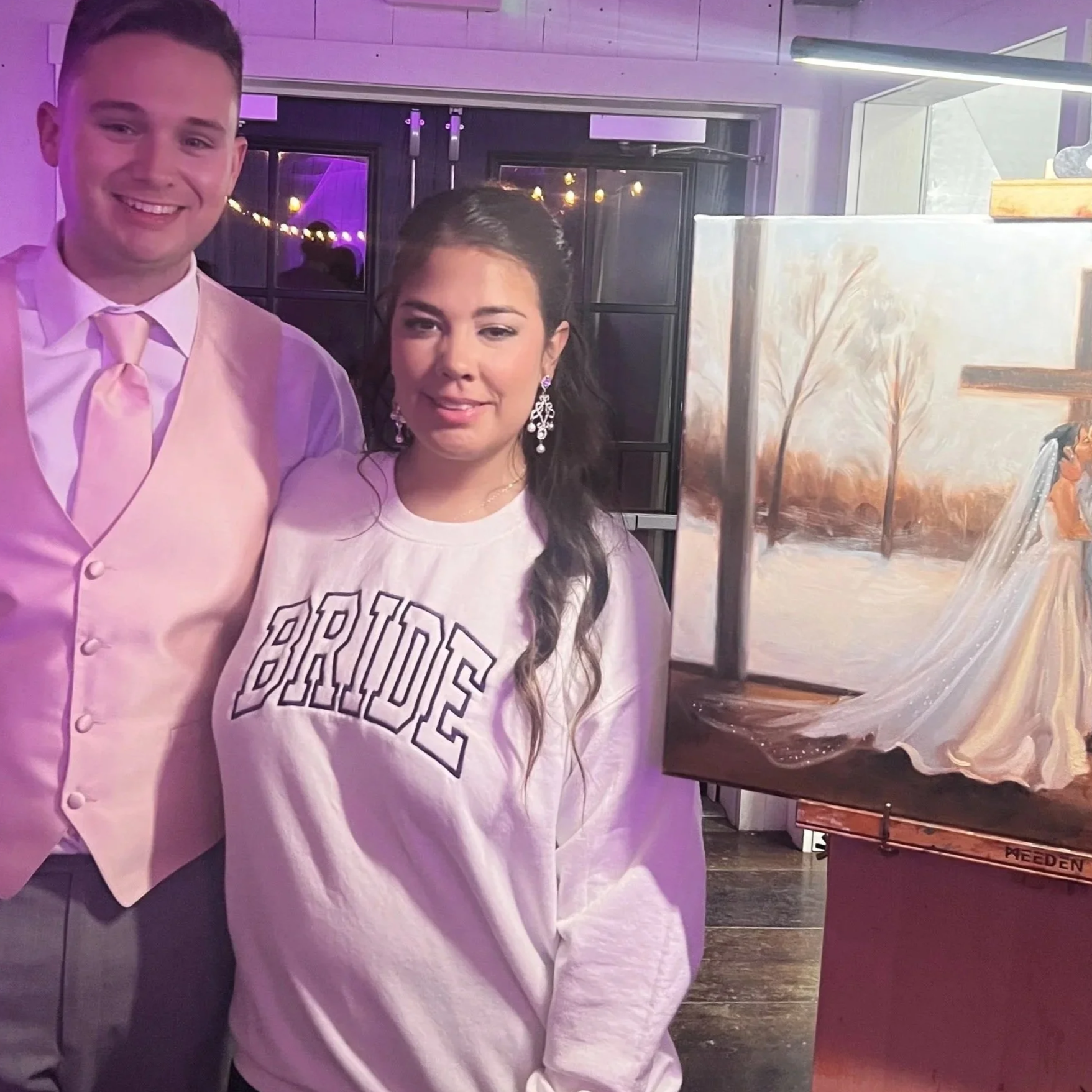 Two people are standing in front of a framed painting and a glass window at an indoor event. The man on the left is smiling, wearing a light pink vest, dress shirt, and tie. The woman on the right is smiling, with long dark hair, wearing large earrings and a light-colored sweatshirt
