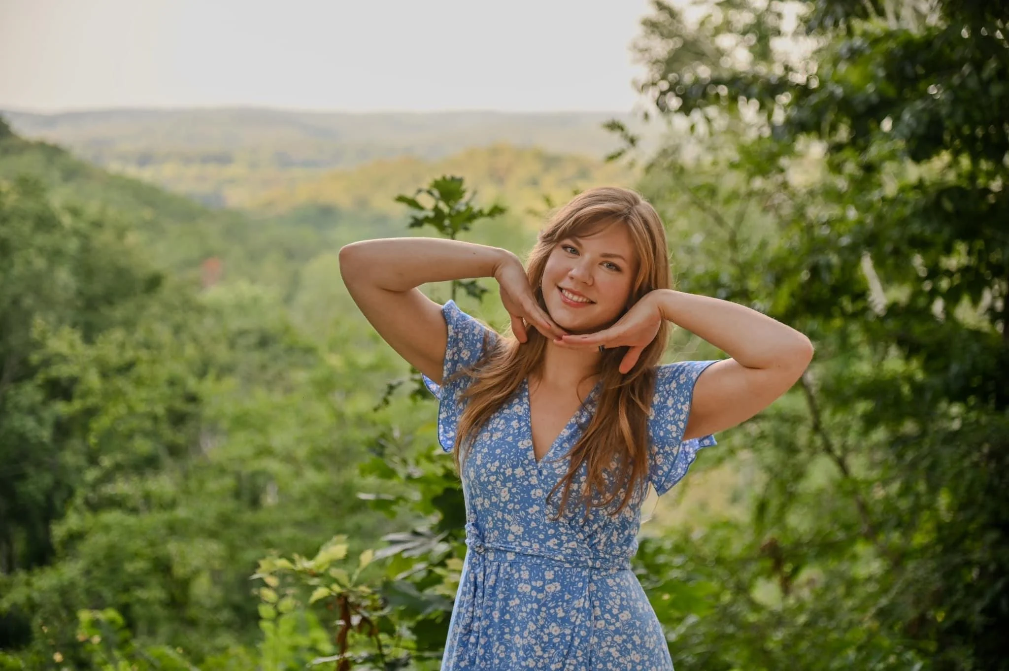 Woman with long red hair, smiling, posing outdoors in a blue floral dress with greenery and trees in the background.