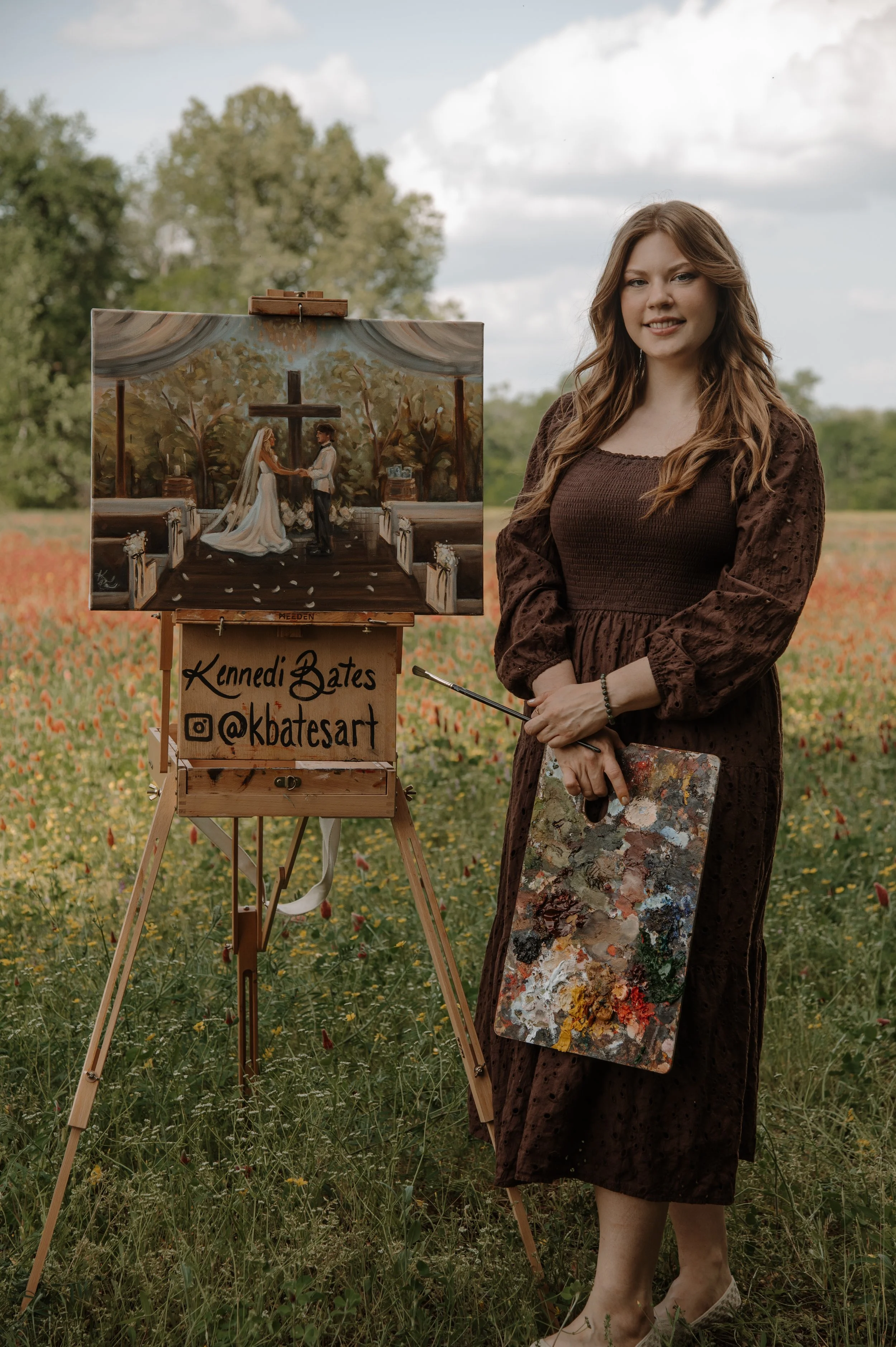 A young woman with long hair in a brown dress stands outdoors in a field of yellow and orange wildflowers, holding a painter's palette and brush, smiling. Beside her is an easel with a painting of a wedding scene inside a church with a cross and a bride and groom holding hands.