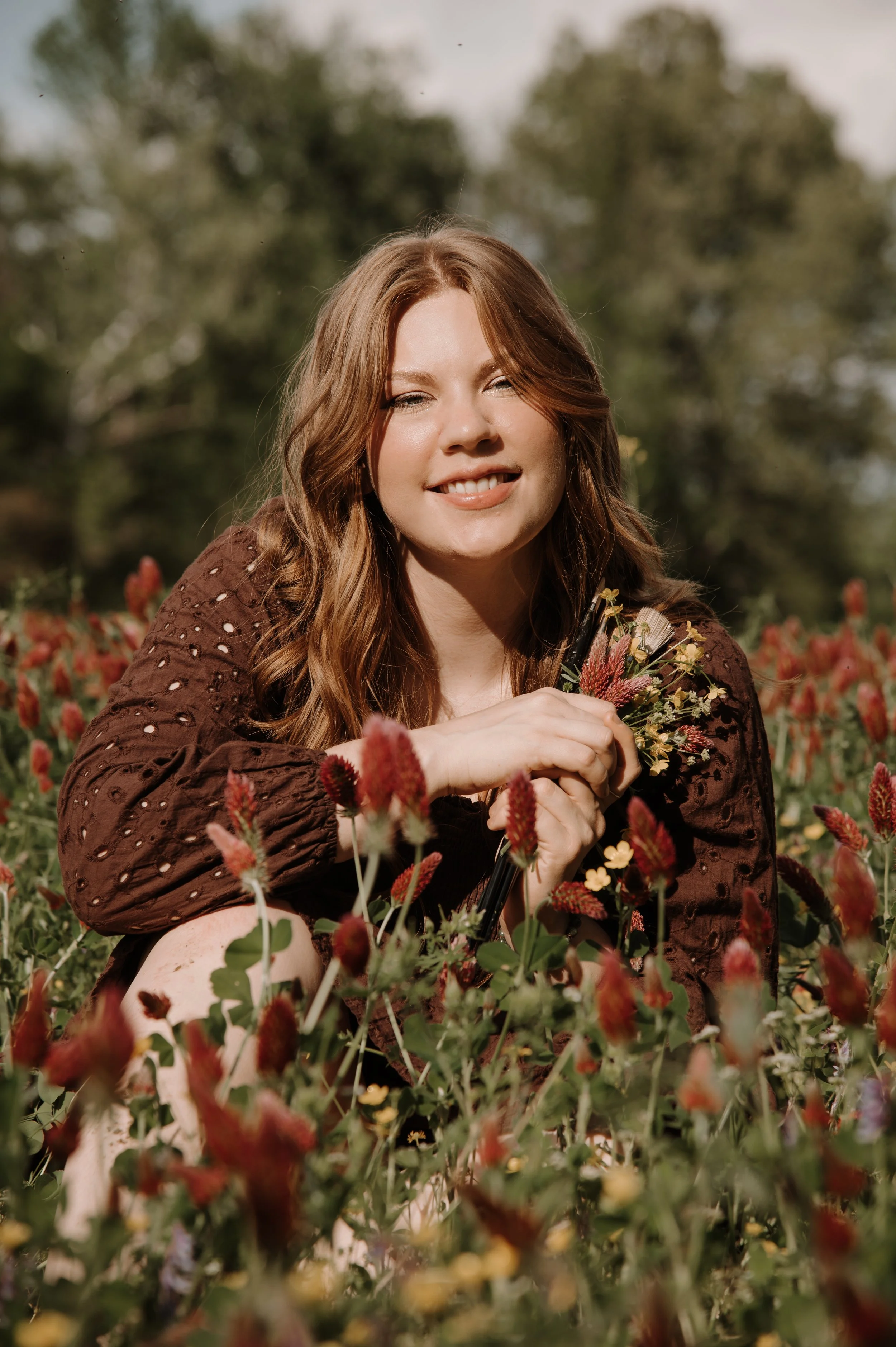 A young woman with long red hair sitting among flowers outdoors, smiling at the camera.