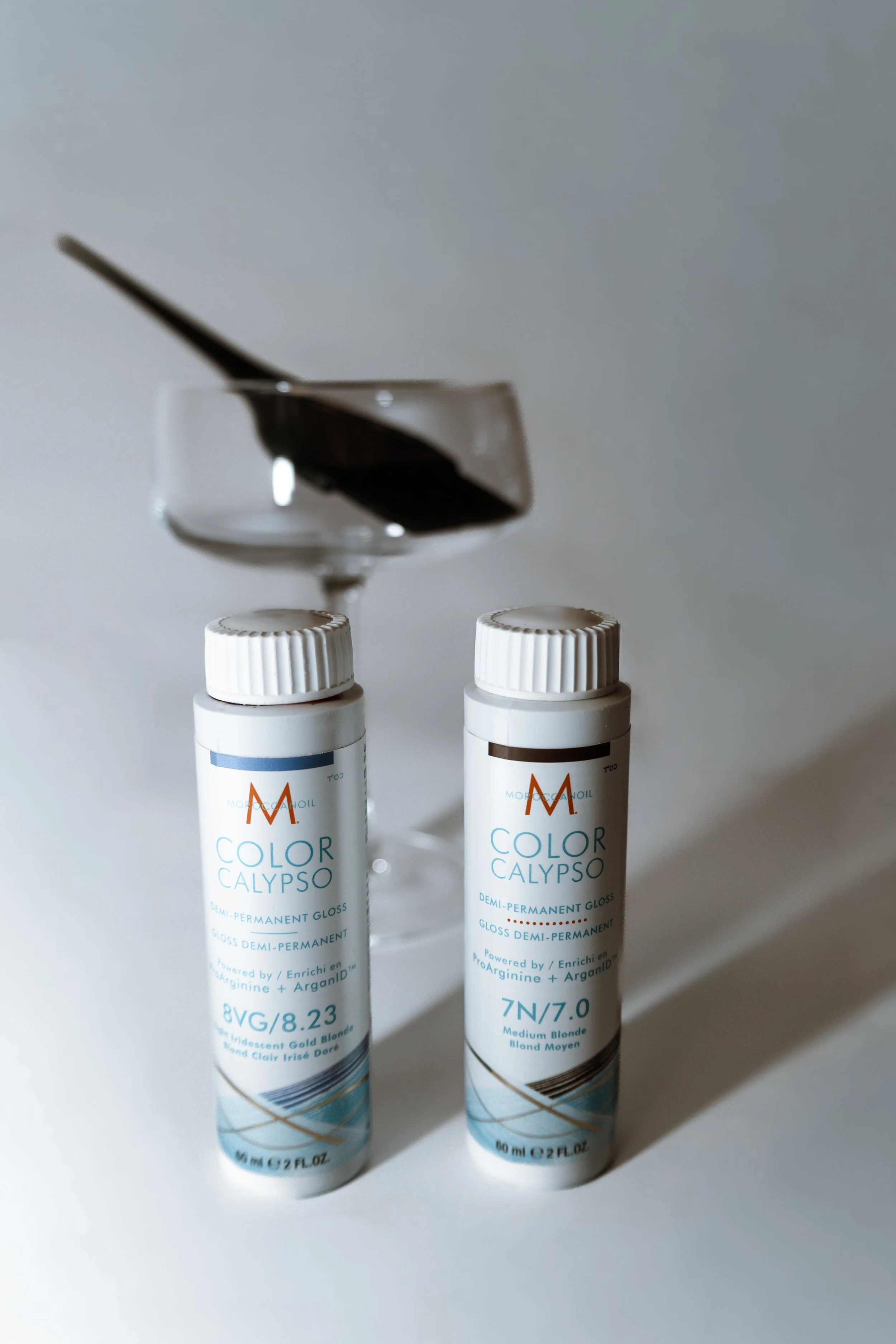Two bottles of semi-permanent hair gloss color Calypso stand on a white surface, with a glass of black hair dye and a brush on top.