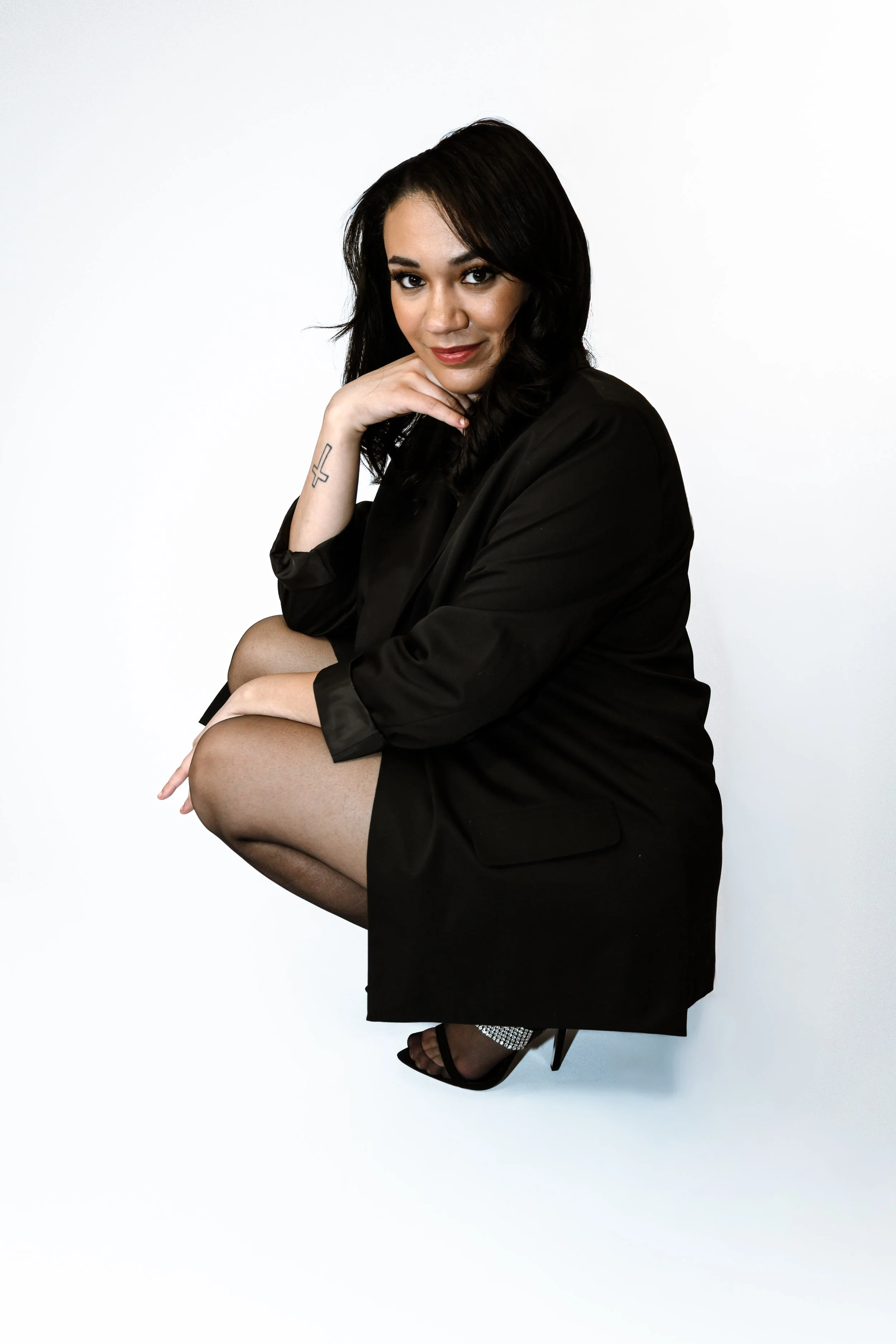 A woman dressed in a black blazer, black stockings, and high heels, kneeling with one knee up and resting her chin on her hand, against a plain white background.