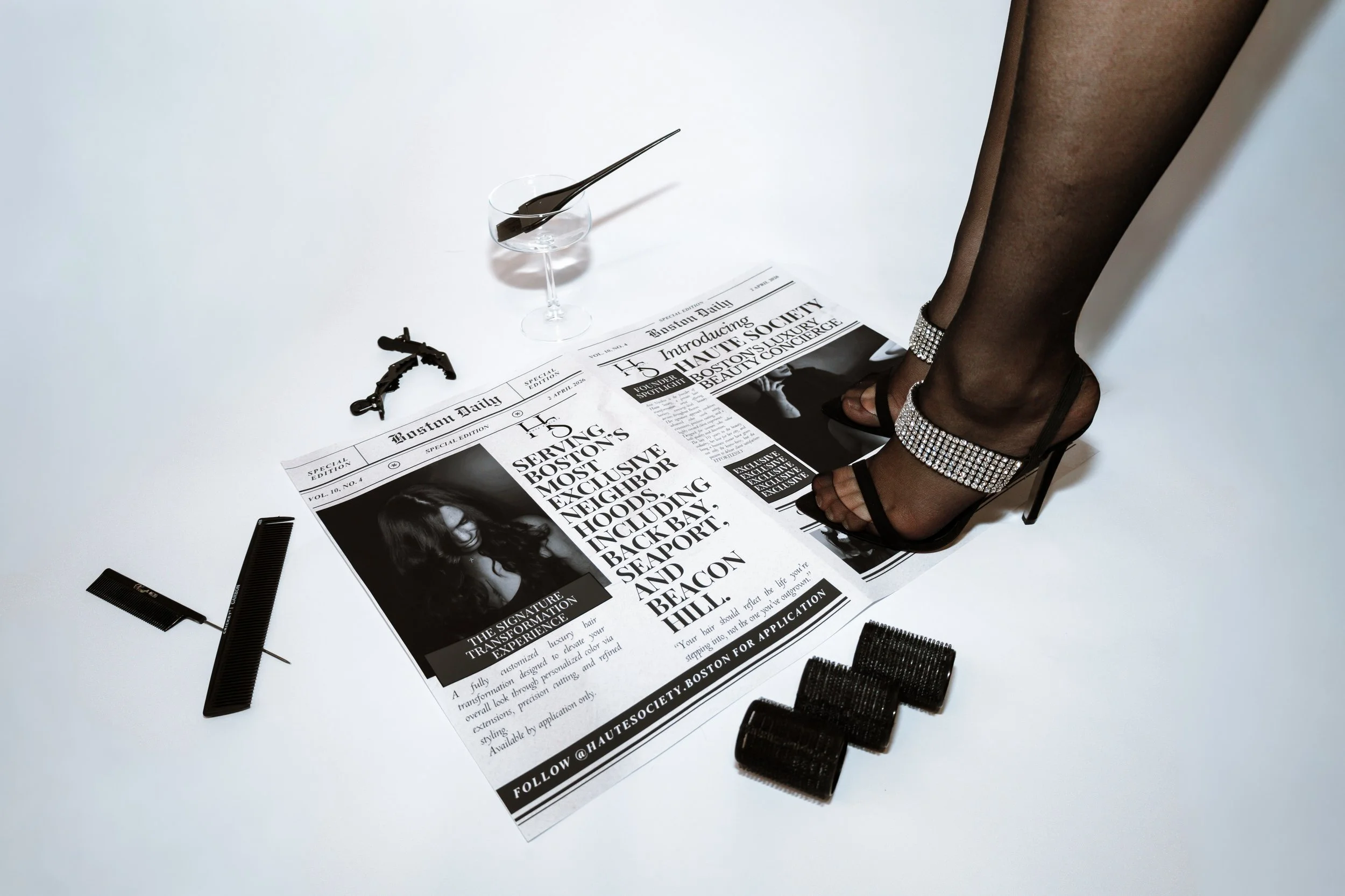 A person's legs in high-heeled shoes with ankle straps decorated with rhinestones are standing on a black and white newspaper on a white surface. Hair styling tools including a comb, hair clips, and hair rollers, along with a small dish with a hairbrush, are also on the newspaper.