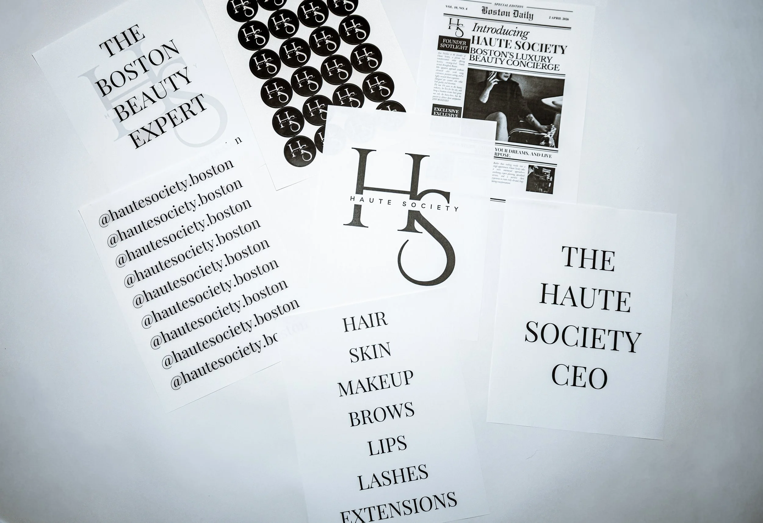 A collection of printed materials, including flyers and cards, related to HAUTE SOCIETY, a beauty and fashion brand. The materials feature the brand's logo, social media handles (@hautesociety.boston), and a list of beauty services such as hair, skin, makeup, brows, lips, lashes, extensions, and more. Some materials include black circular stickers with the brand's logo.