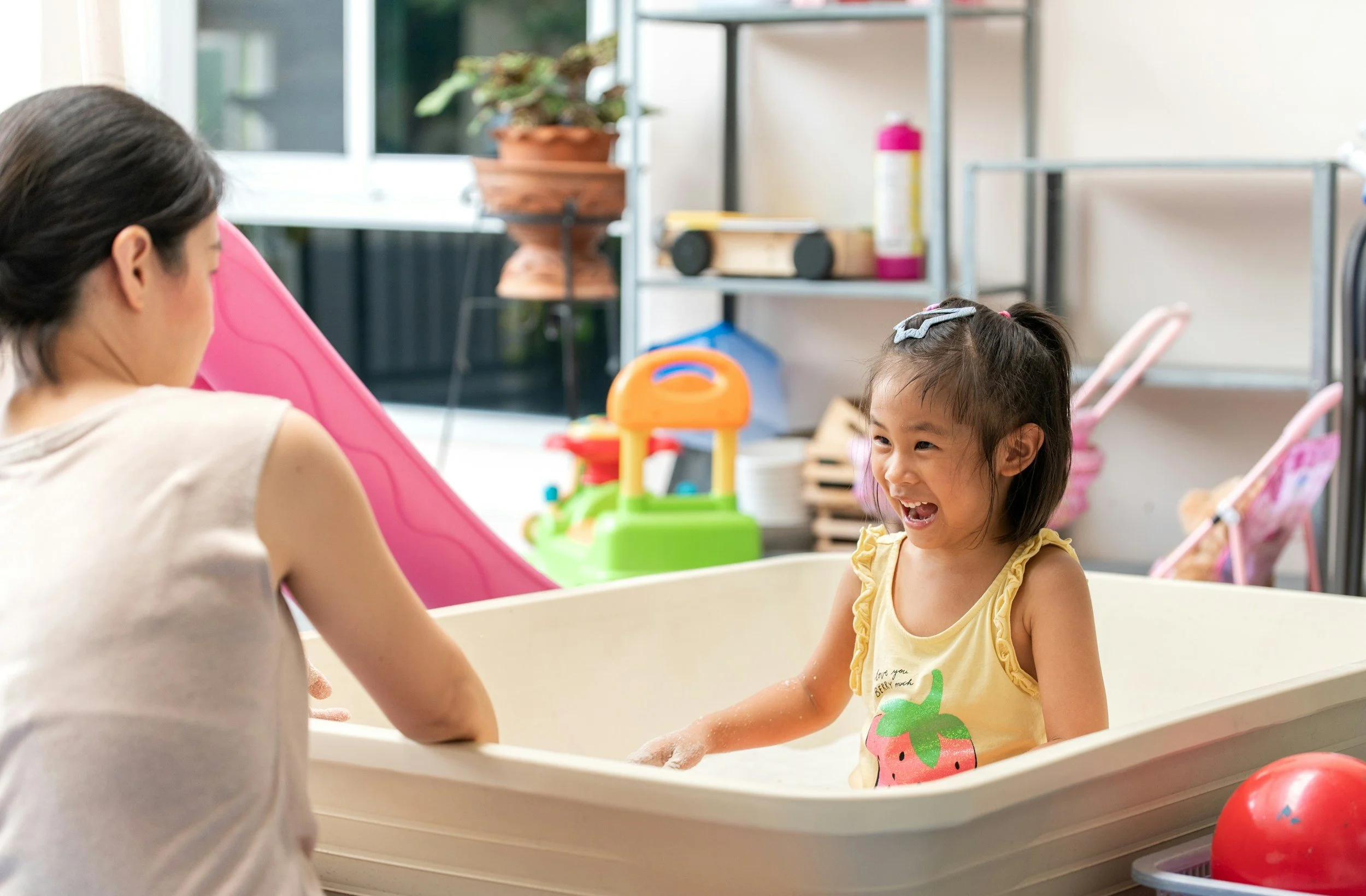 A young girl with a gray bow in her hair is sitting in a small plastic tub, smiling and laughing while interacting with an adult woman who is also in the tub. The scene takes place indoors, with a play area and toys visible in the background.