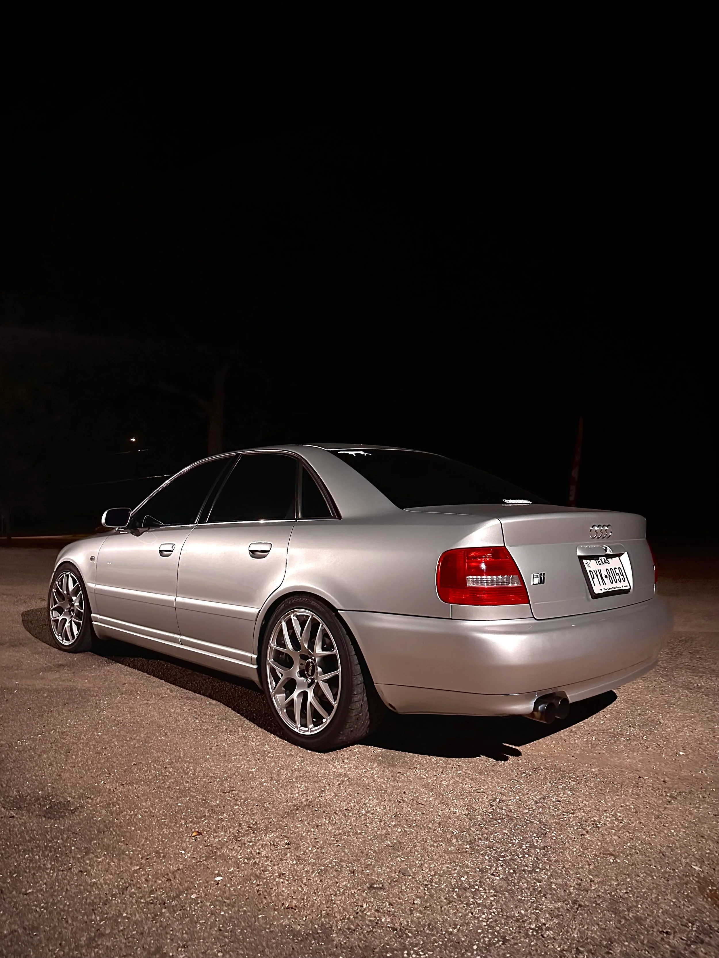 A silver Audi sedan parked on a dirt surface at night, with a dark sky and few trees in the background.