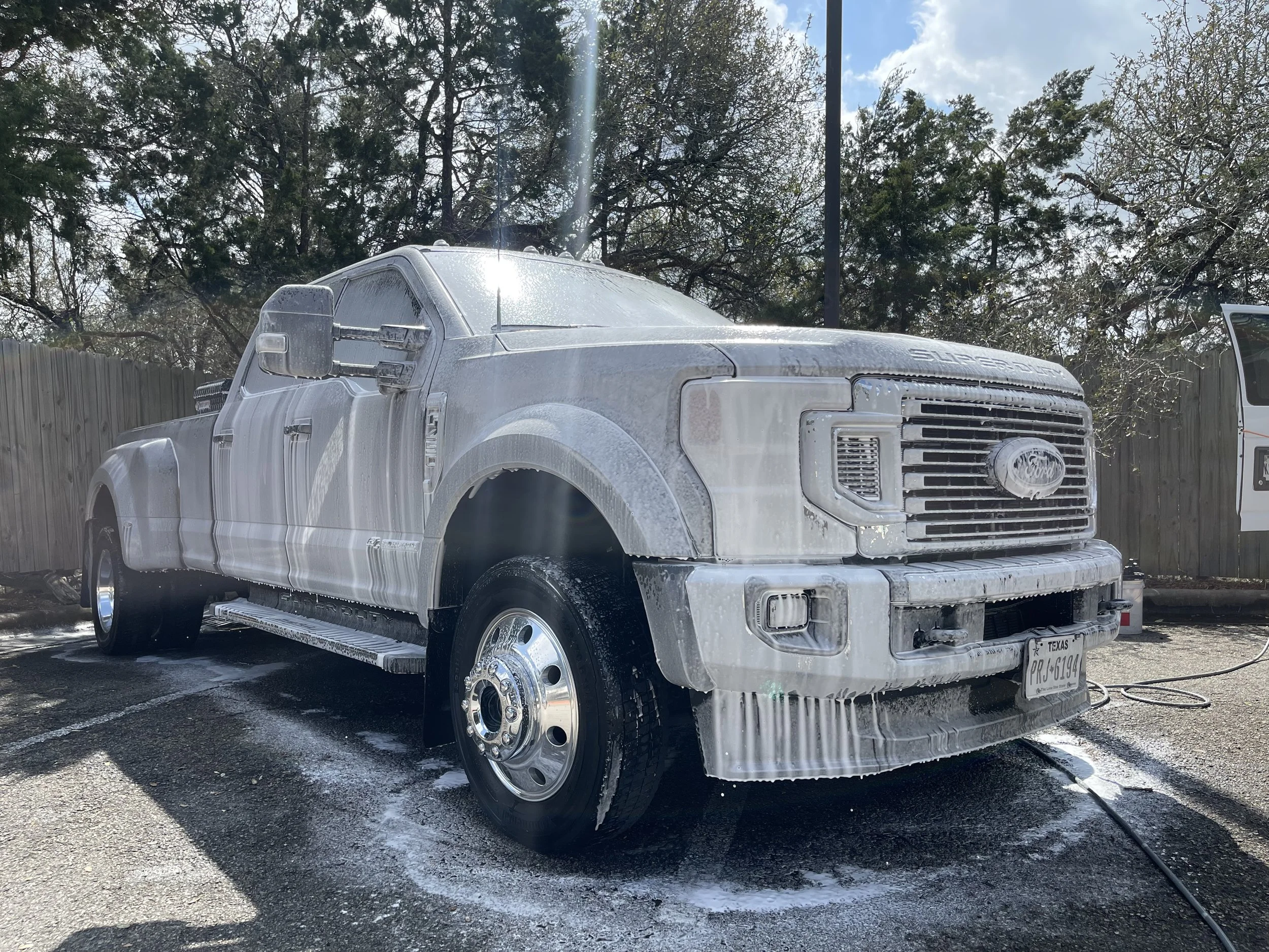 A large Ford pickup truck covered in white foam during a car wash in a parking lot. The foam covers the entire truck, and soap suds are visible on the ground around the tires.