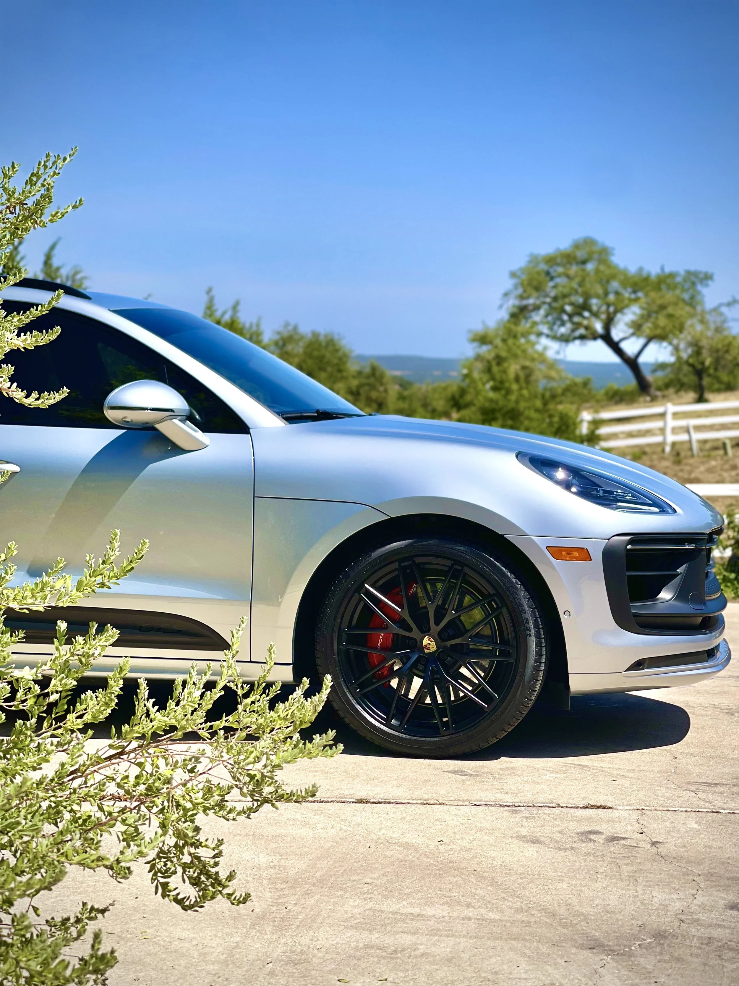 A silver sports car parked on a driveway with green bushes in the foreground, trees, a white fence, and a clear blue sky in the background.