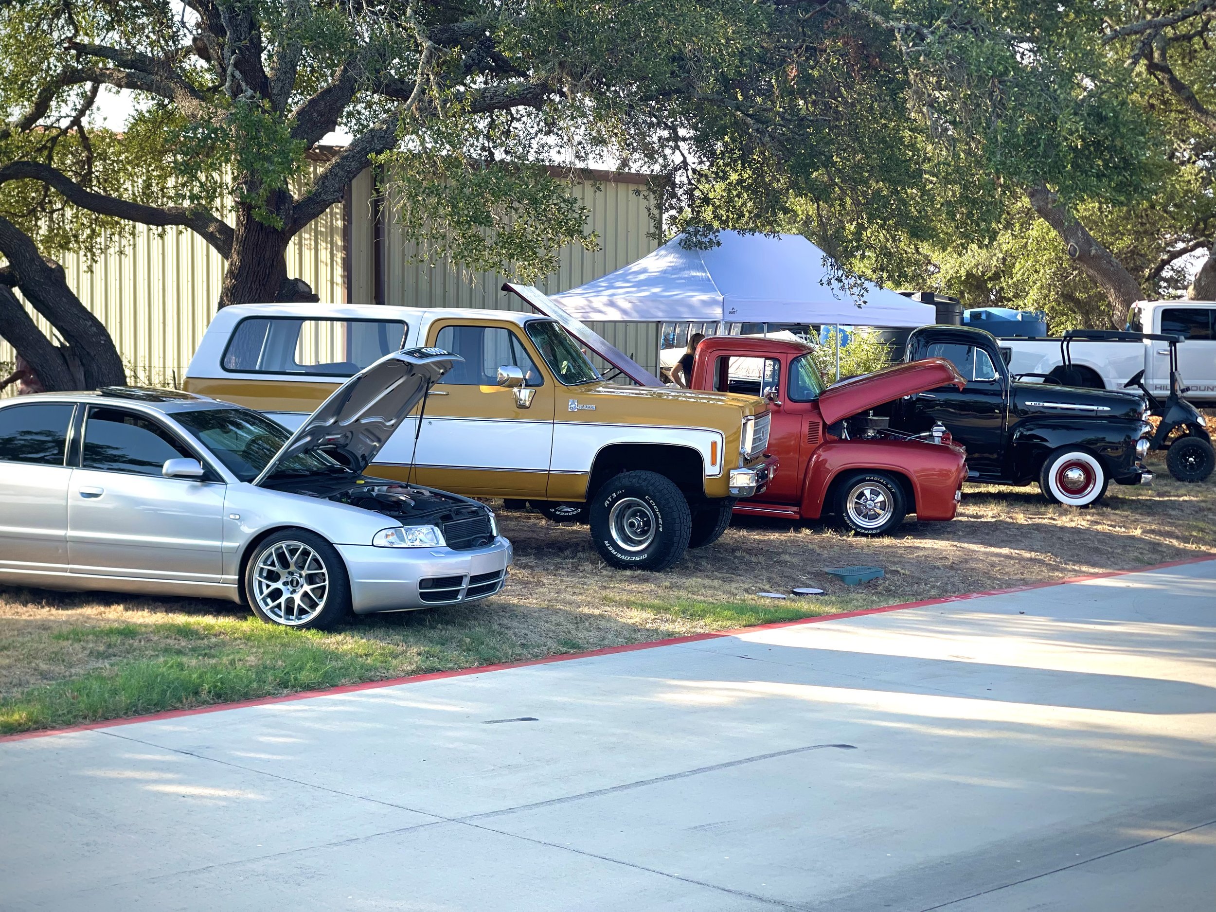 Five vehicles parked outdoors under a tree, with four open hoods revealing engines. There is a silver sedan, a vintage white and gold SUV, a red pickup truck, a black truck, and a golf cart, with a white canopy tent in the background.