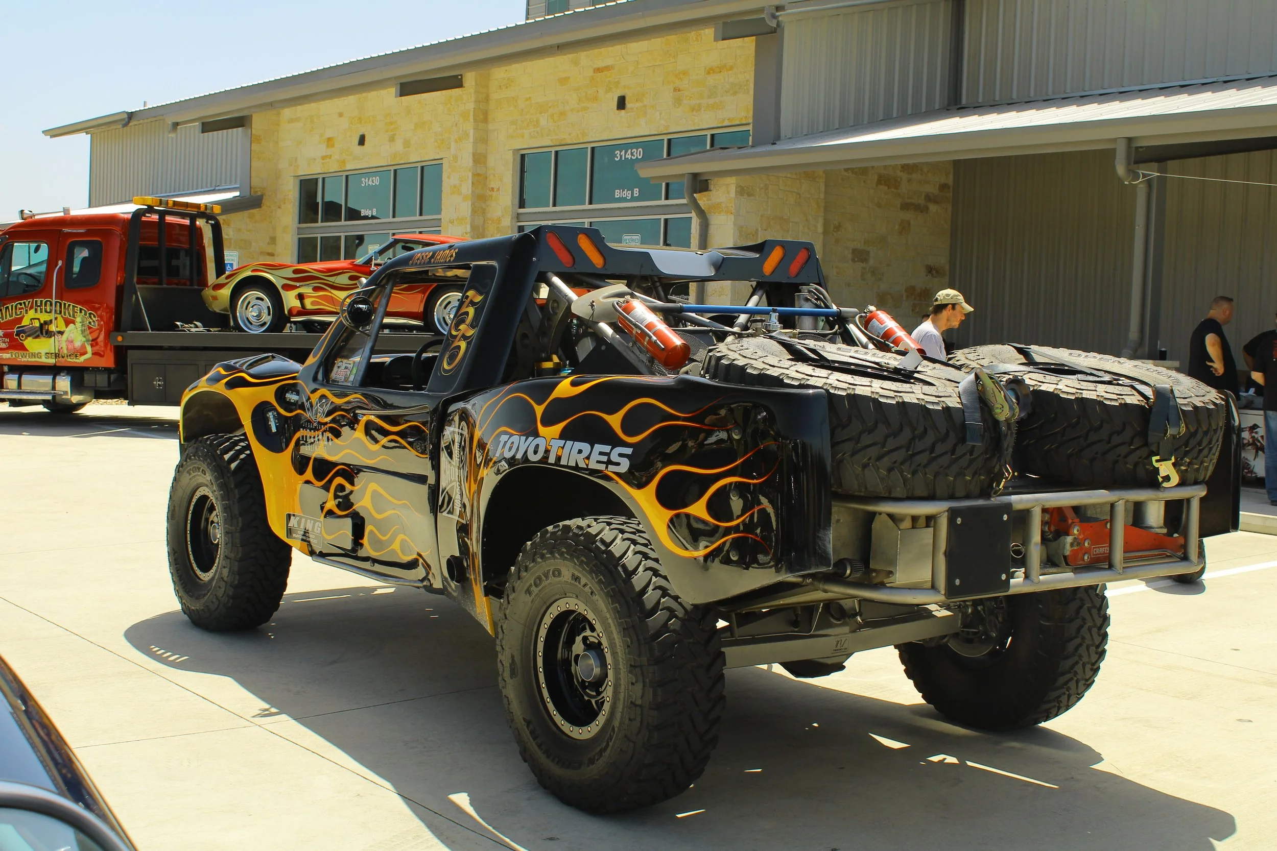 A black off-road racing pickup truck with flame decals and large tires, parked outdoors near a building with people in the background.