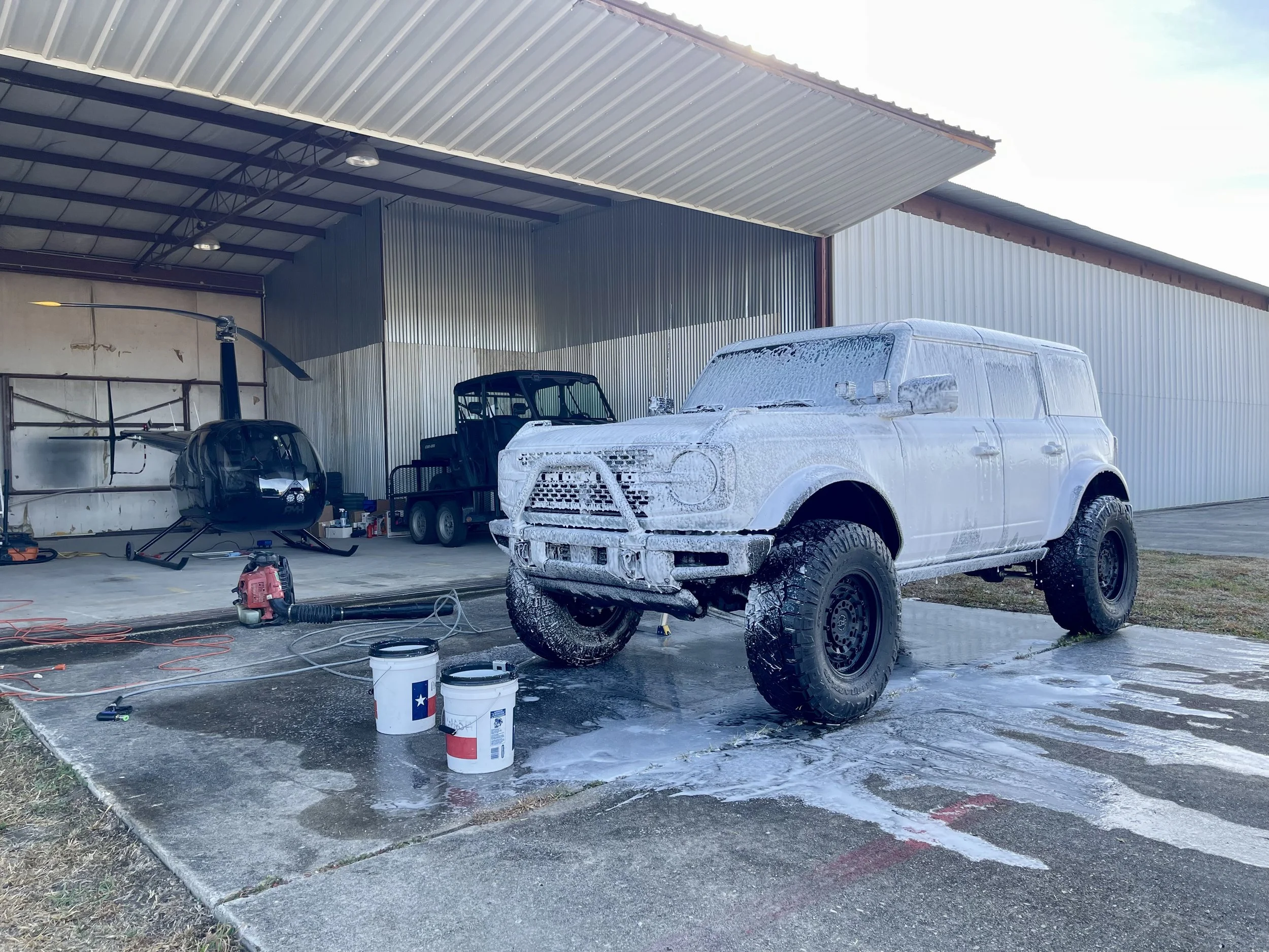 A white off-road vehicle being washed with soap and foam, parked outside a metal garage. There is a black helicopter, a small black utility cart, a water pump, and cleaning supplies nearby.
