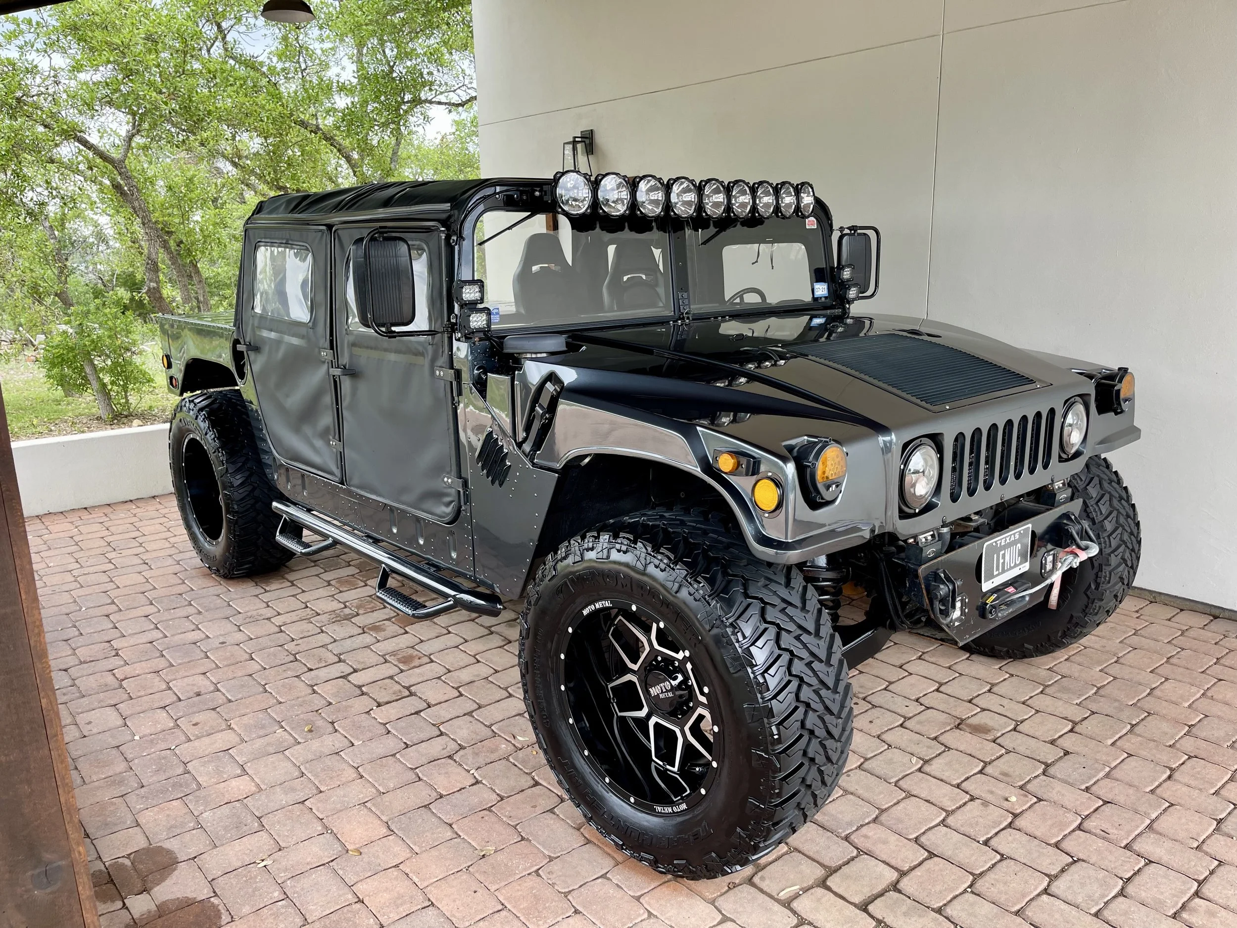 Black modified off-road Jeep with large tires, top row of lights, and a Texas license plate reading 'LFMIC', parked on a brick patio with greenery and trees in the background.
