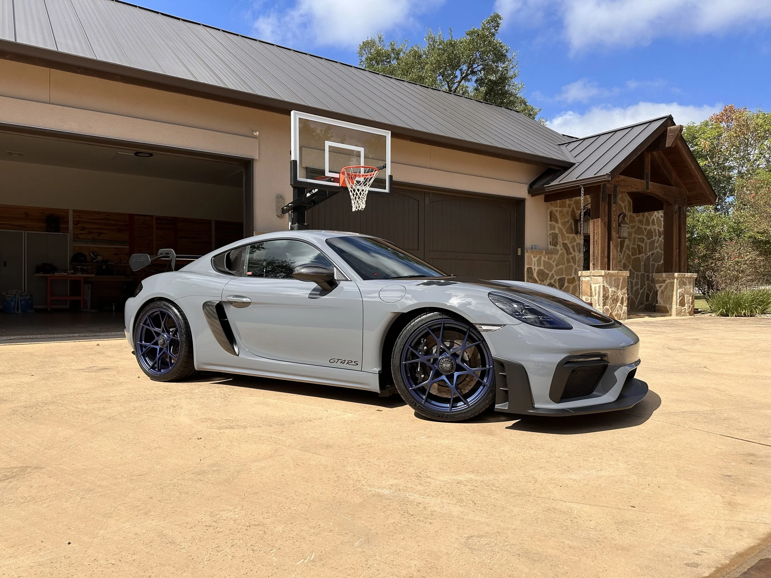 A silver sports car parked outside a house with an open garage and a basketball hoop attached to the house, on a sunny day.