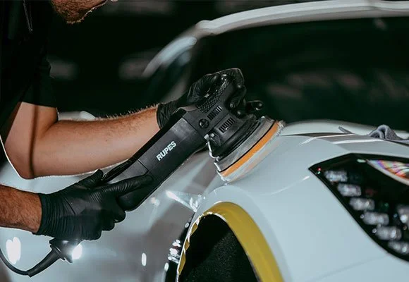 Person using a Rupes polishing machine to buff a car's white paint in a garage.