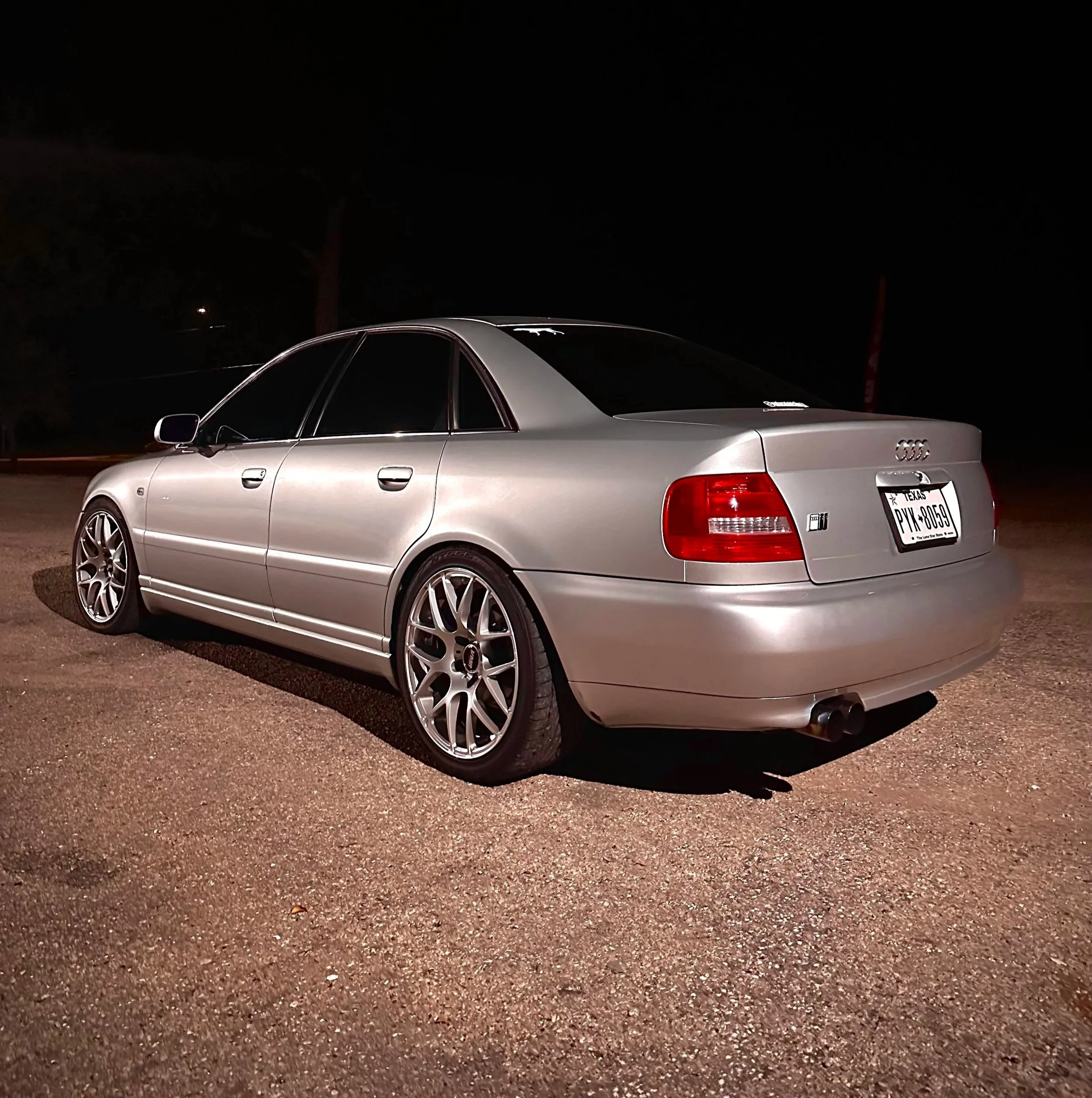 A silver Audi sedan parked on a gravel surface at night, with custom alloy wheels and tinted windows.
