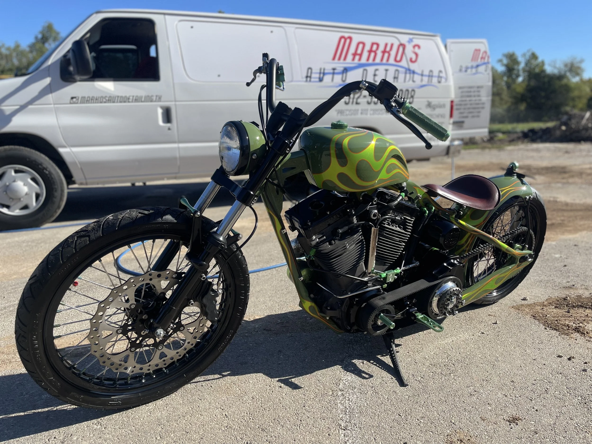 A custom green motorcycle with flames parked outdoors, with a white van in the background advertising auto detailing services.