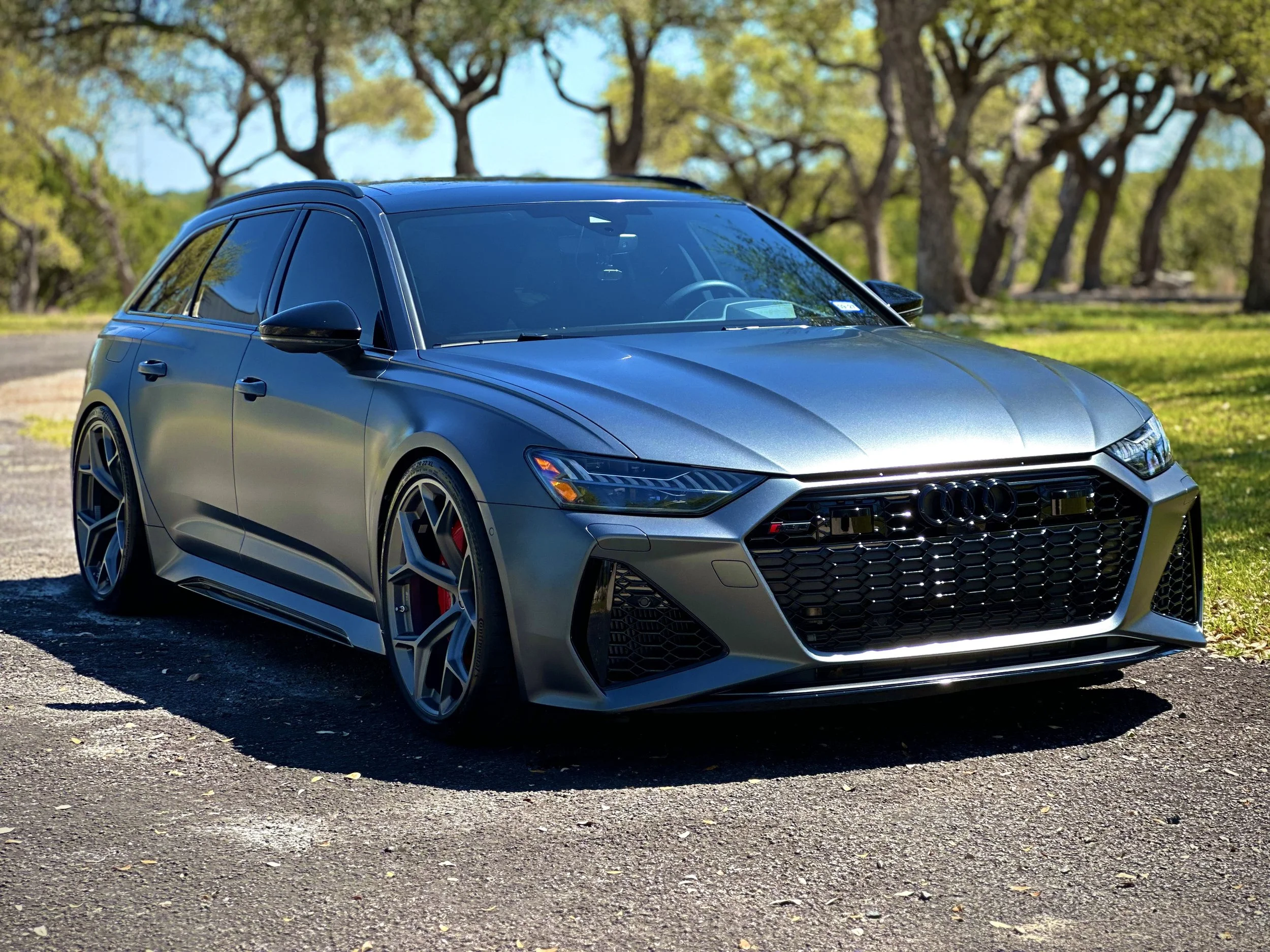 A silver Audi RS series hatchback car parked outdoors on a dirt surface with trees and a blue sky in the background.