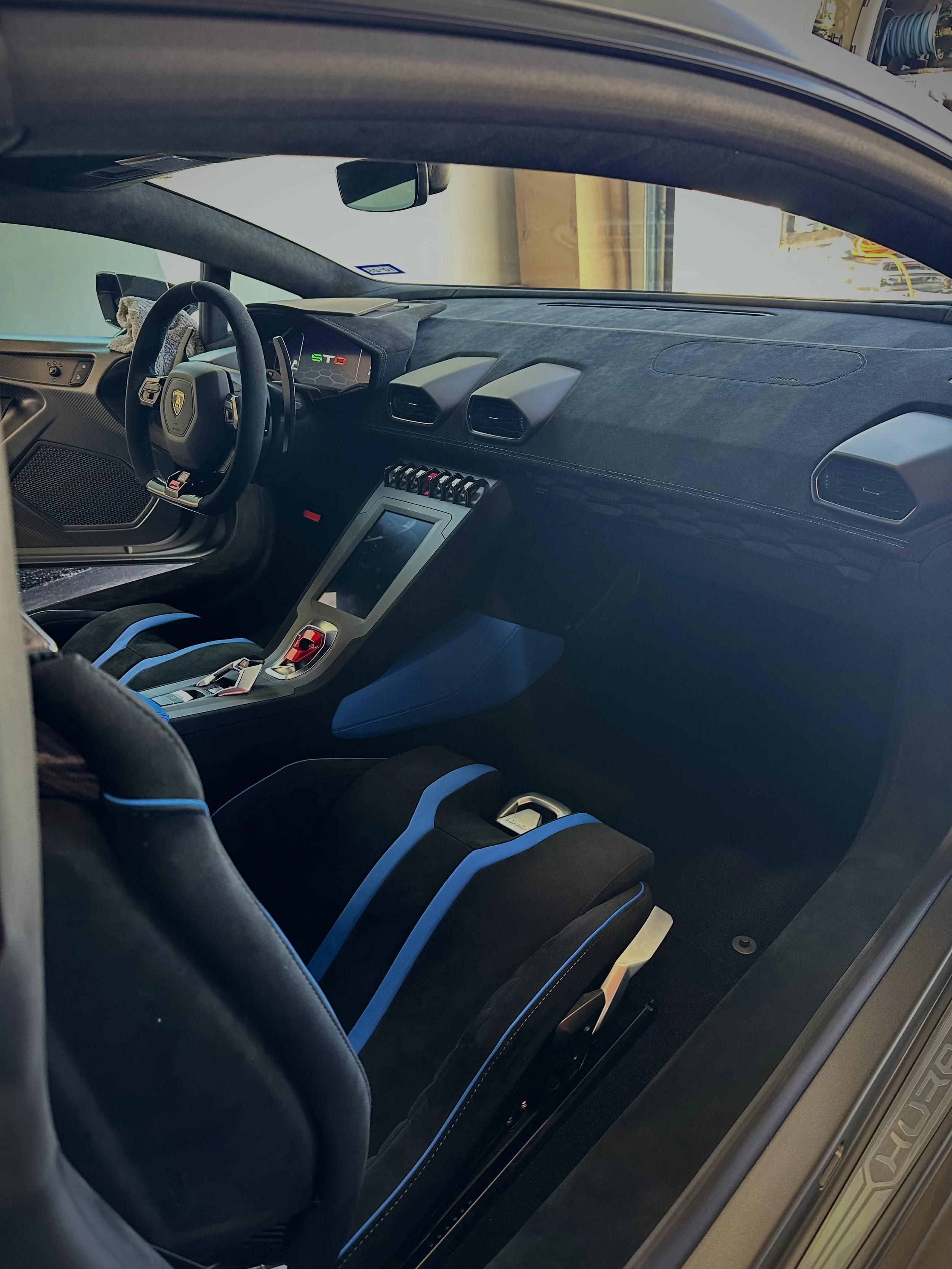 The interior of a sports car, likely a Lamborghini, with a dashboard displaying an 'STO' badge, racing-style seats with blue accents, and a central touchscreen control system.