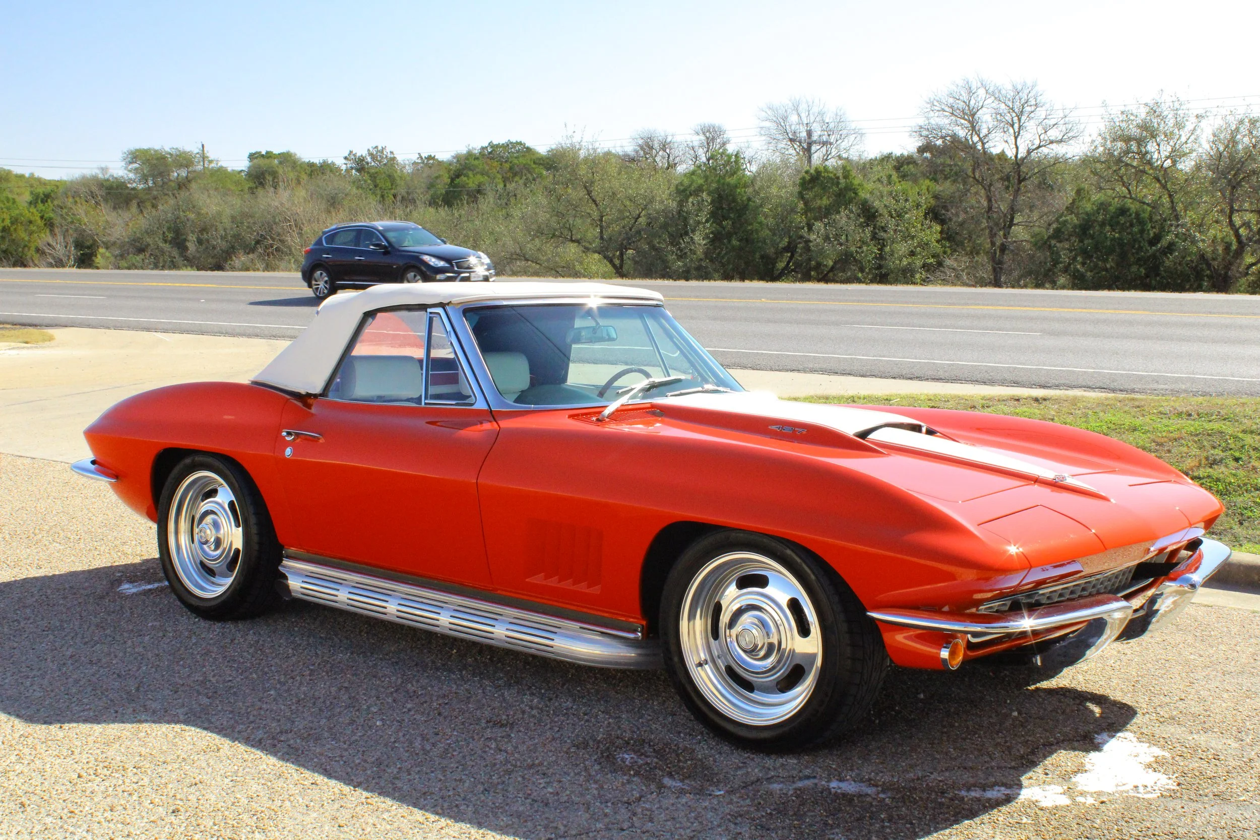 A vintage red Chevrolet Corvette with a white convertible top parked on a roadside, with a black car driving on the road in the background and trees in the distance.