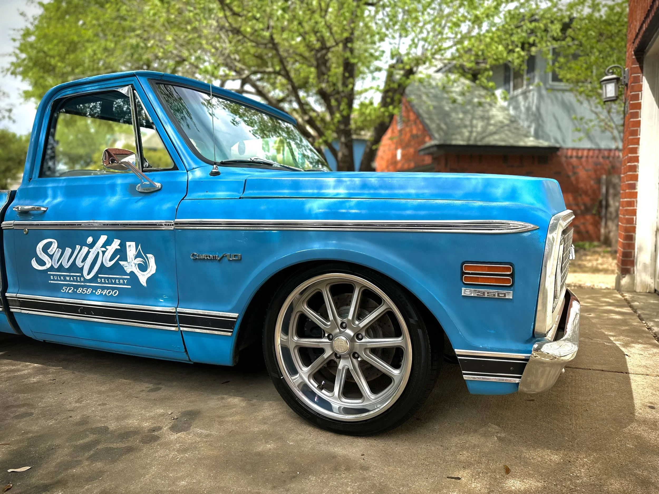 A vintage blue pickup truck with Swift bulk water delivery logo parked on a driveway in front of a red brick house with trees in the background.