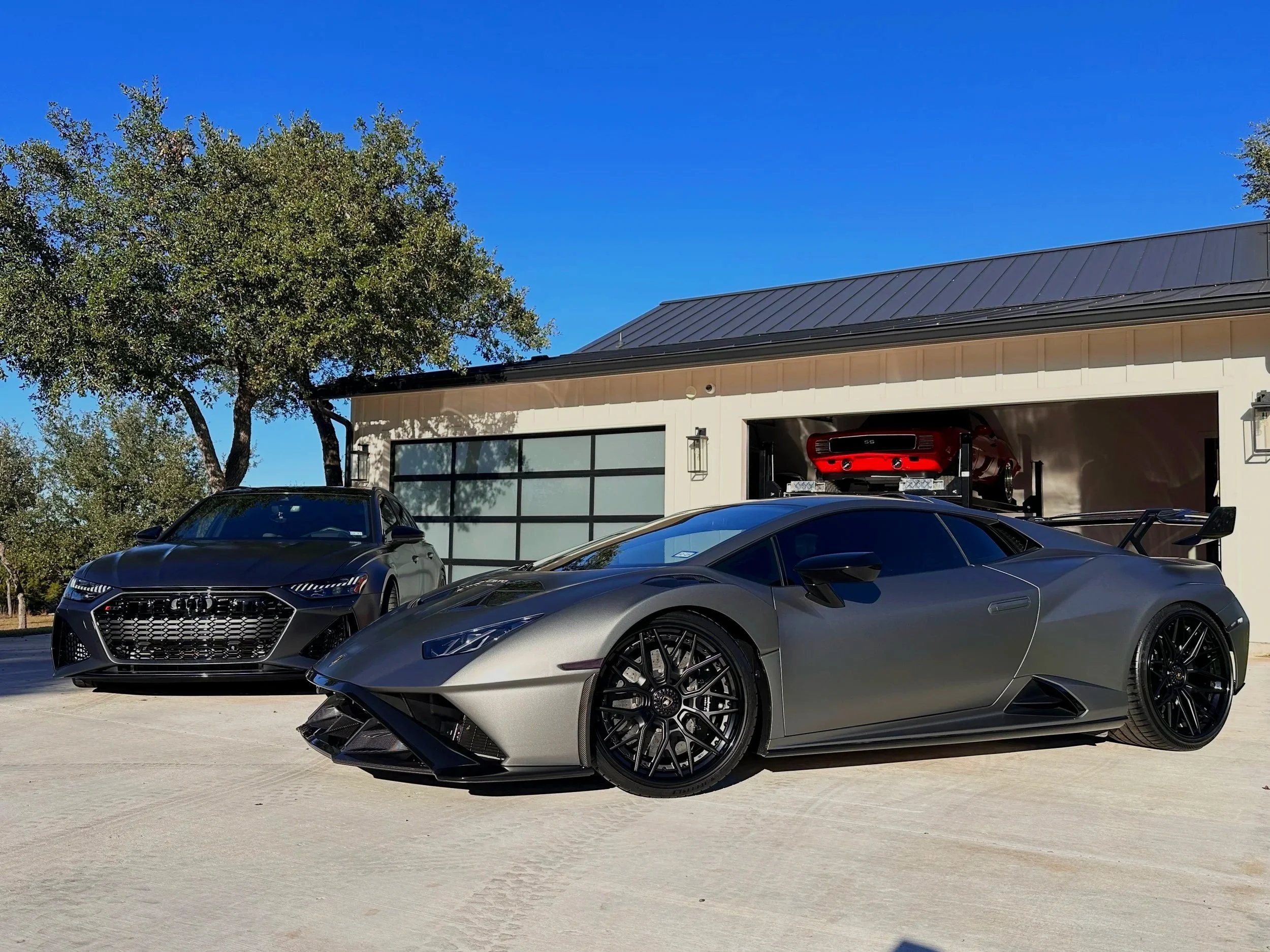 A black Audi sports car and a gray Lamborghini with a black wing are parked in front of a house with a garage, with a red race car inside the garage, under a clear blue sky.