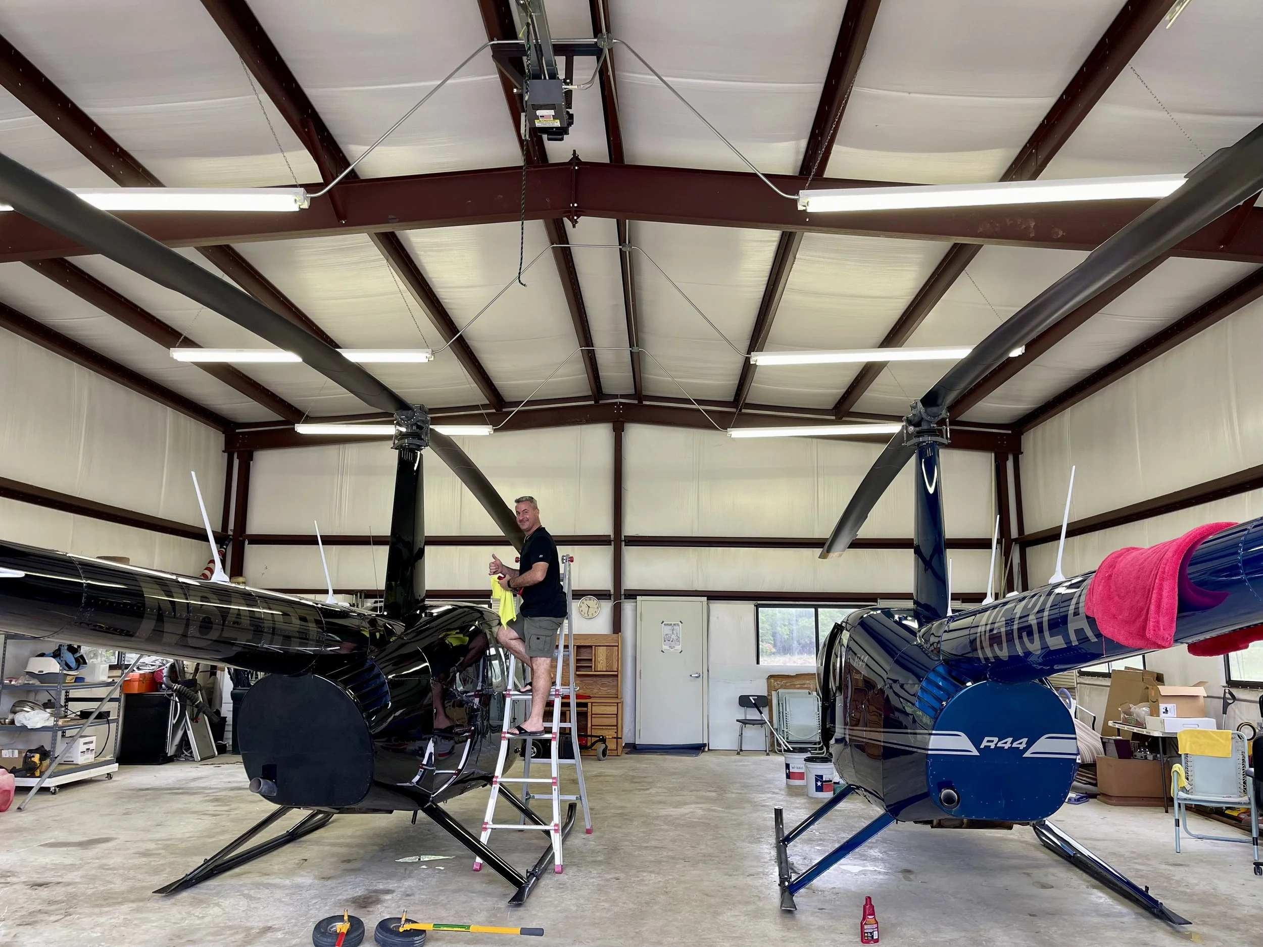 A man standing on a ladder working on two small black helicopters inside a workshop or hangar with a high ceiling, fluorescent lighting, and various tools and equipment around.