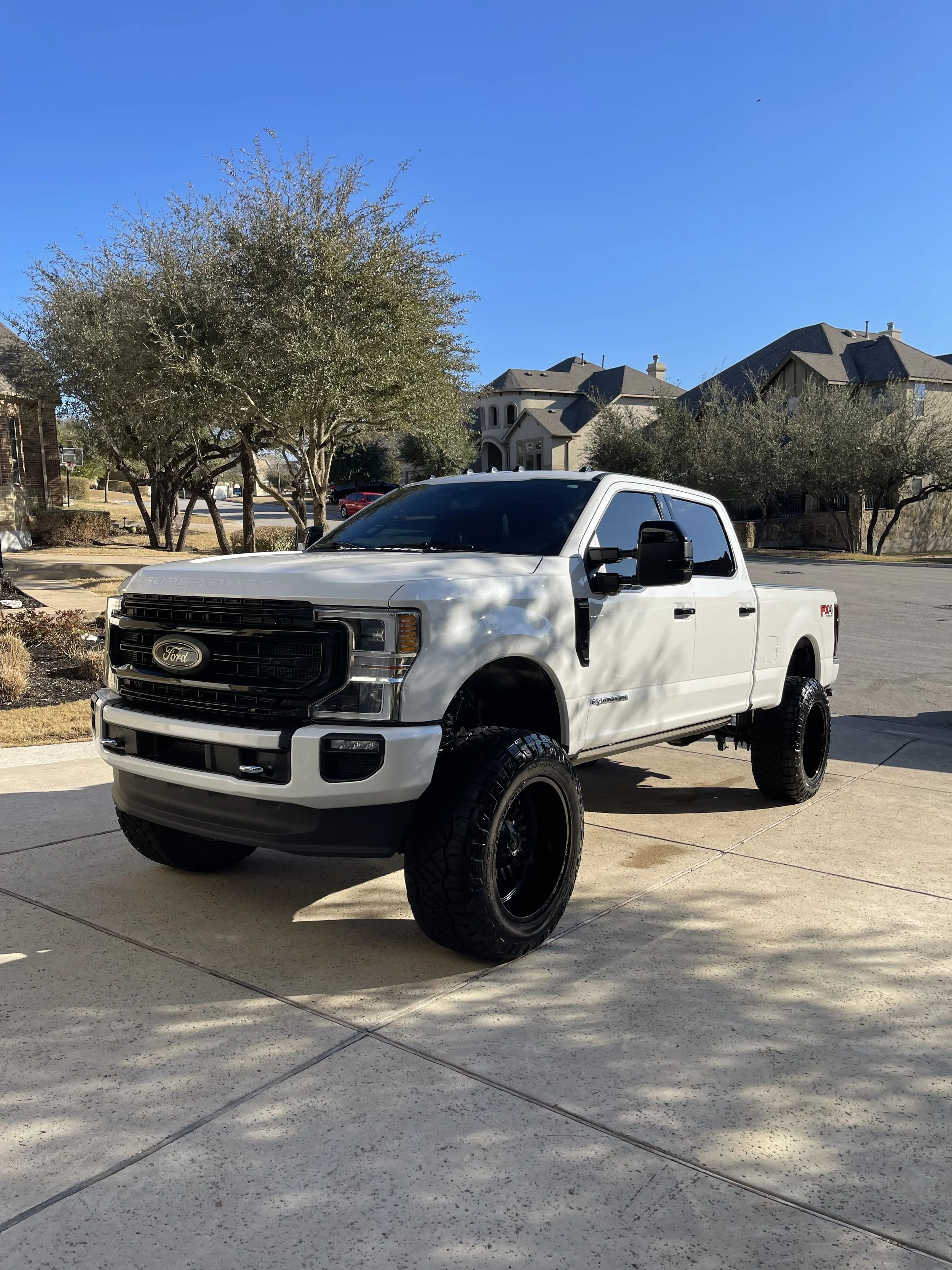White Ford pickup truck with large tires parked on driveway in a suburban neighborhood.