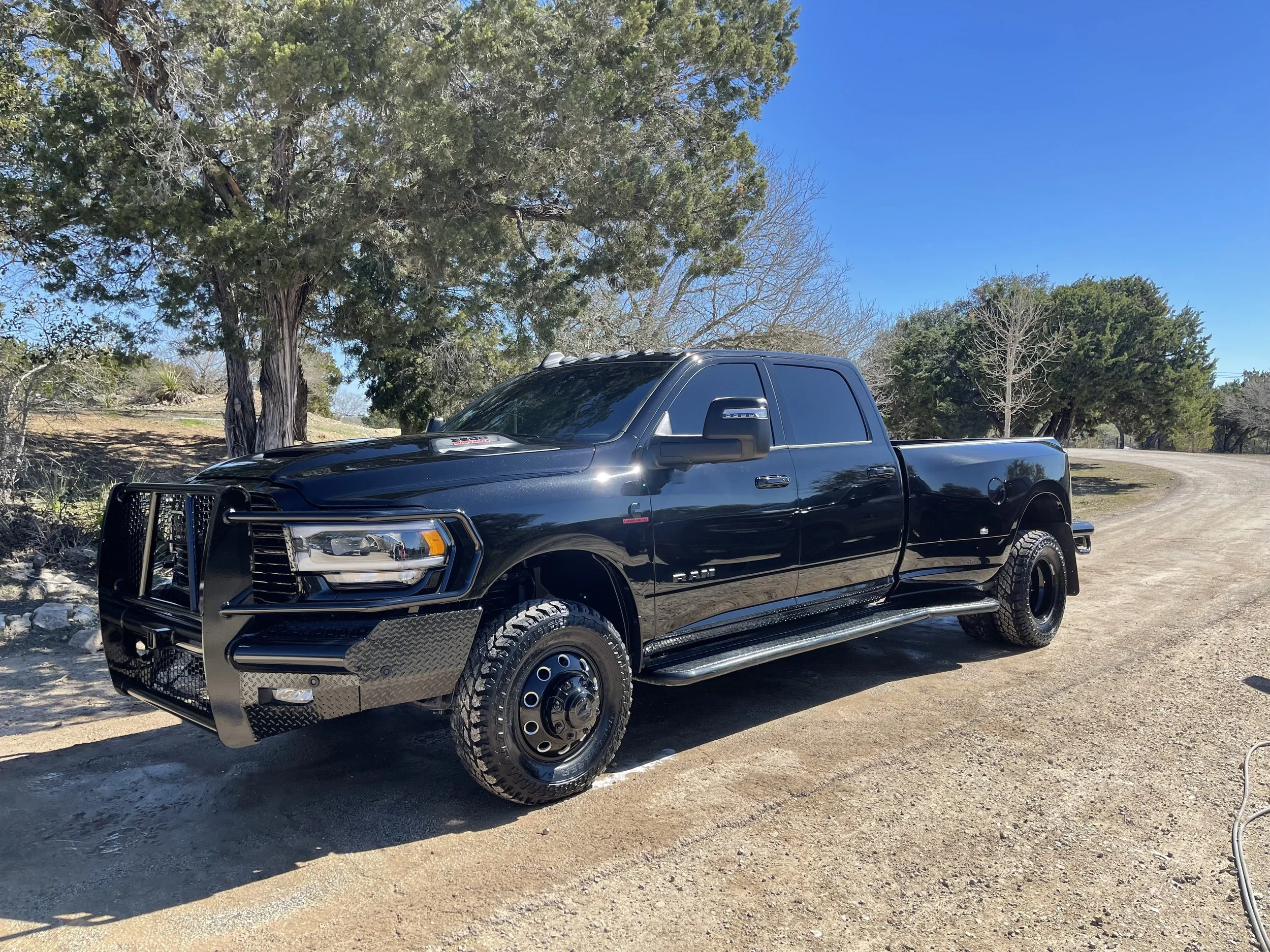 Black pickup truck with a push bar on the front, parked on a dirt road in a rural area with trees and a clear blue sky.