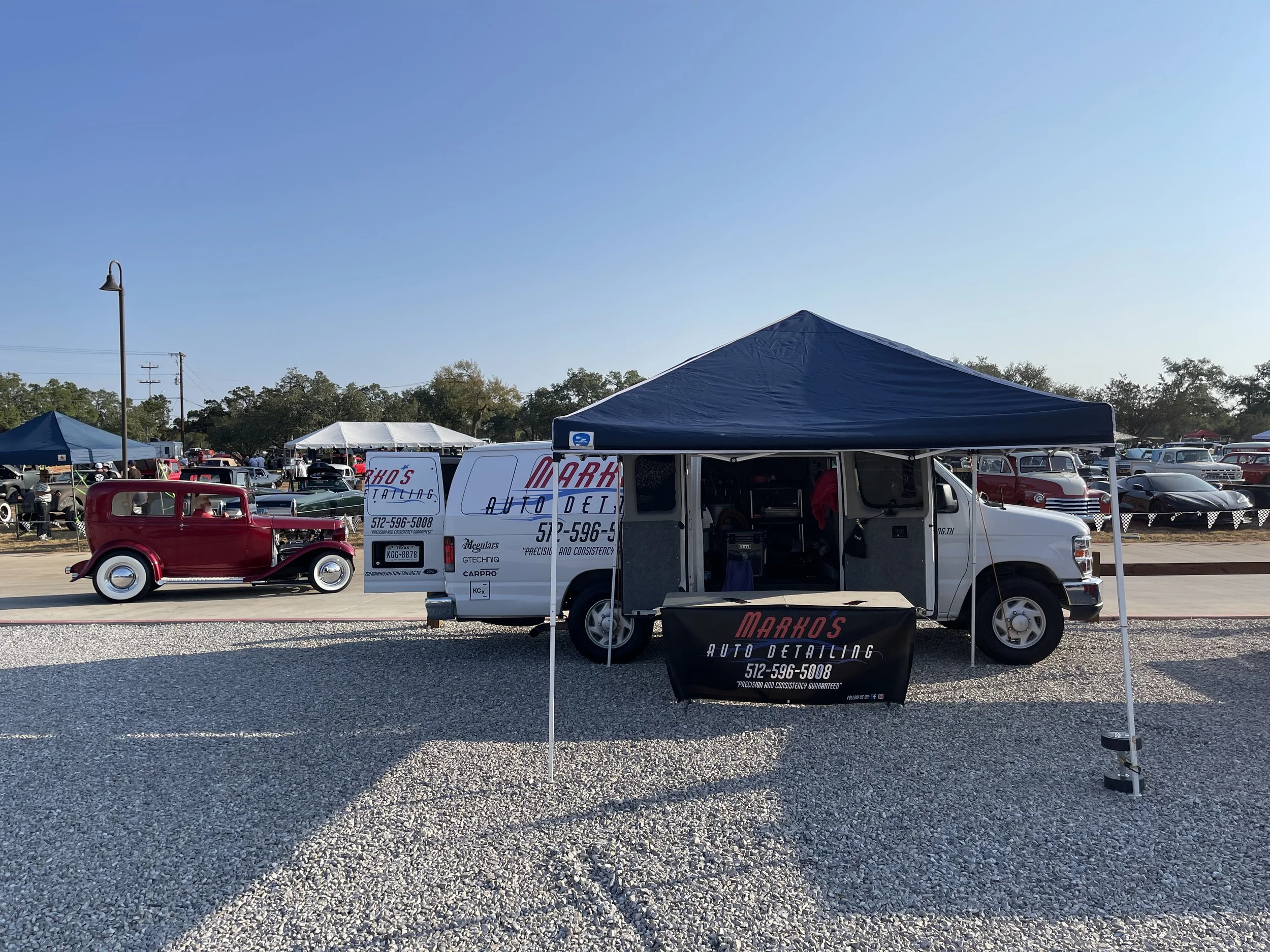 A vintage red car parked next to a white auto detailing van at a classic car show under a clear blue sky. The van has signage for Markos Auto Detailing with a contact number, and a black tent is set up around the vehicle.