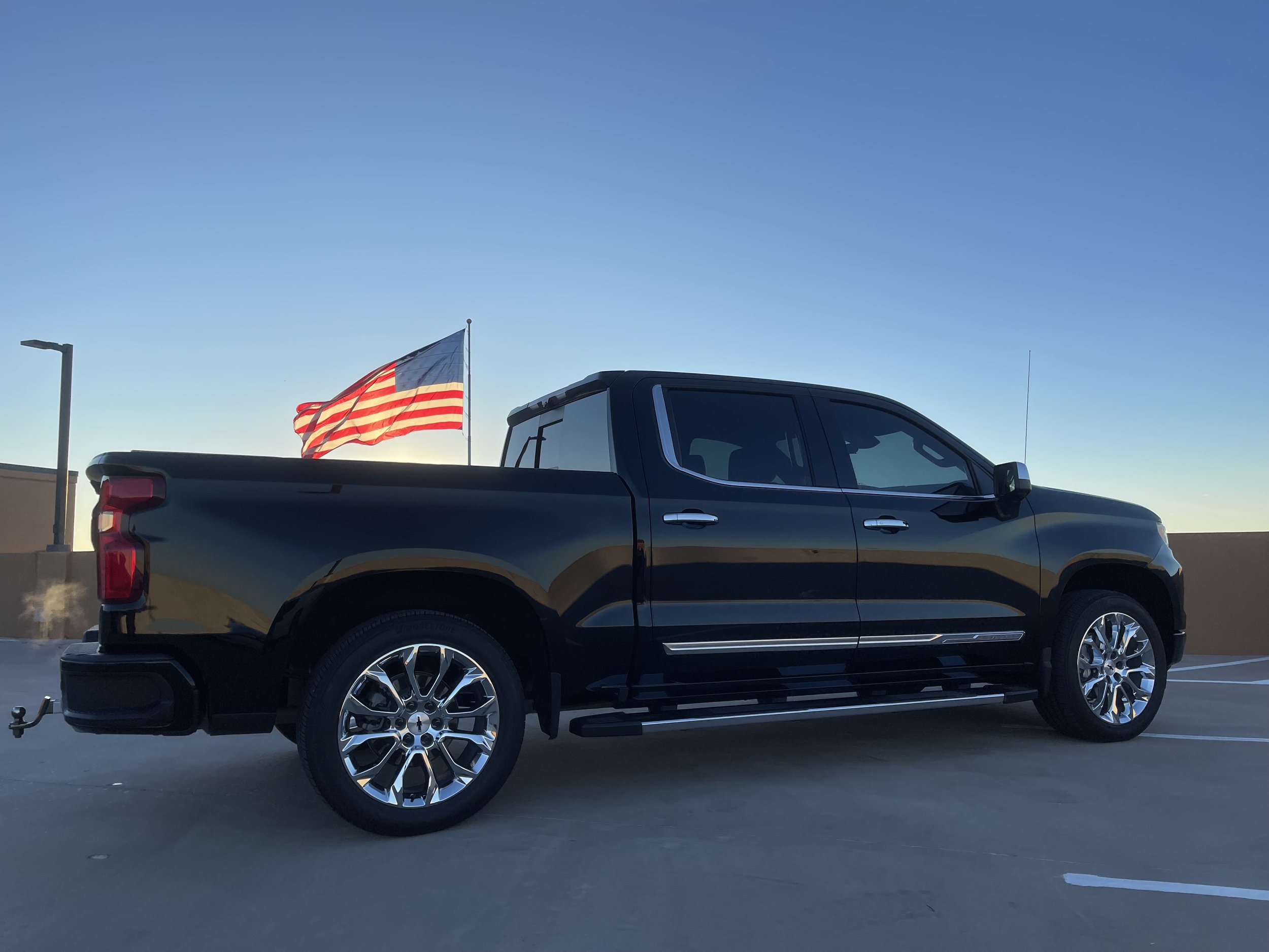 Black pickup truck parked on a rooftop parking lot with an American flag waving in the background during dusk.