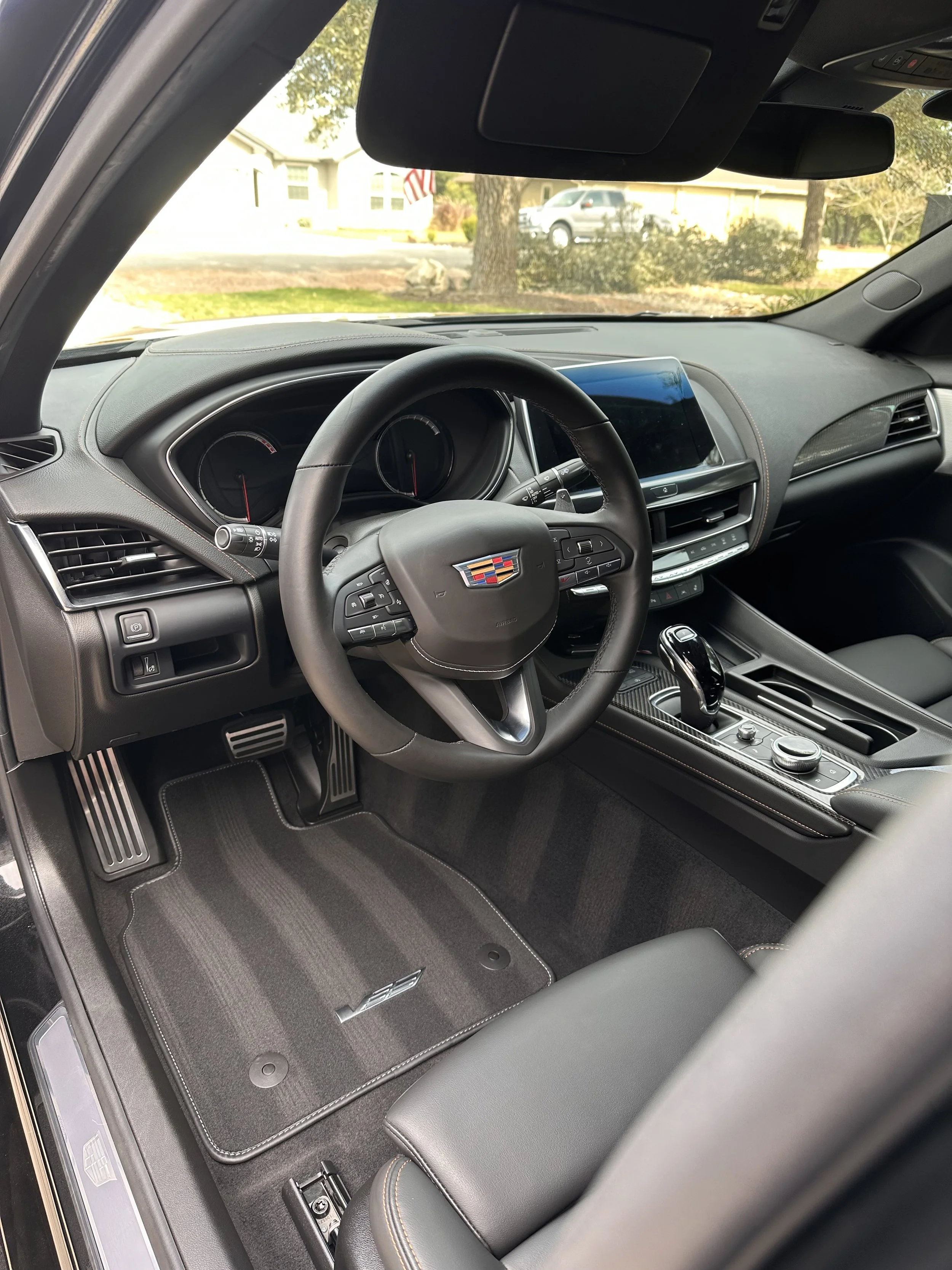 Interior of a Cadillac vehicle showing the steering wheel, dashboard, center console with gear shifter, and a view of the front windshield looking out to a residential neighborhood with houses and trees.