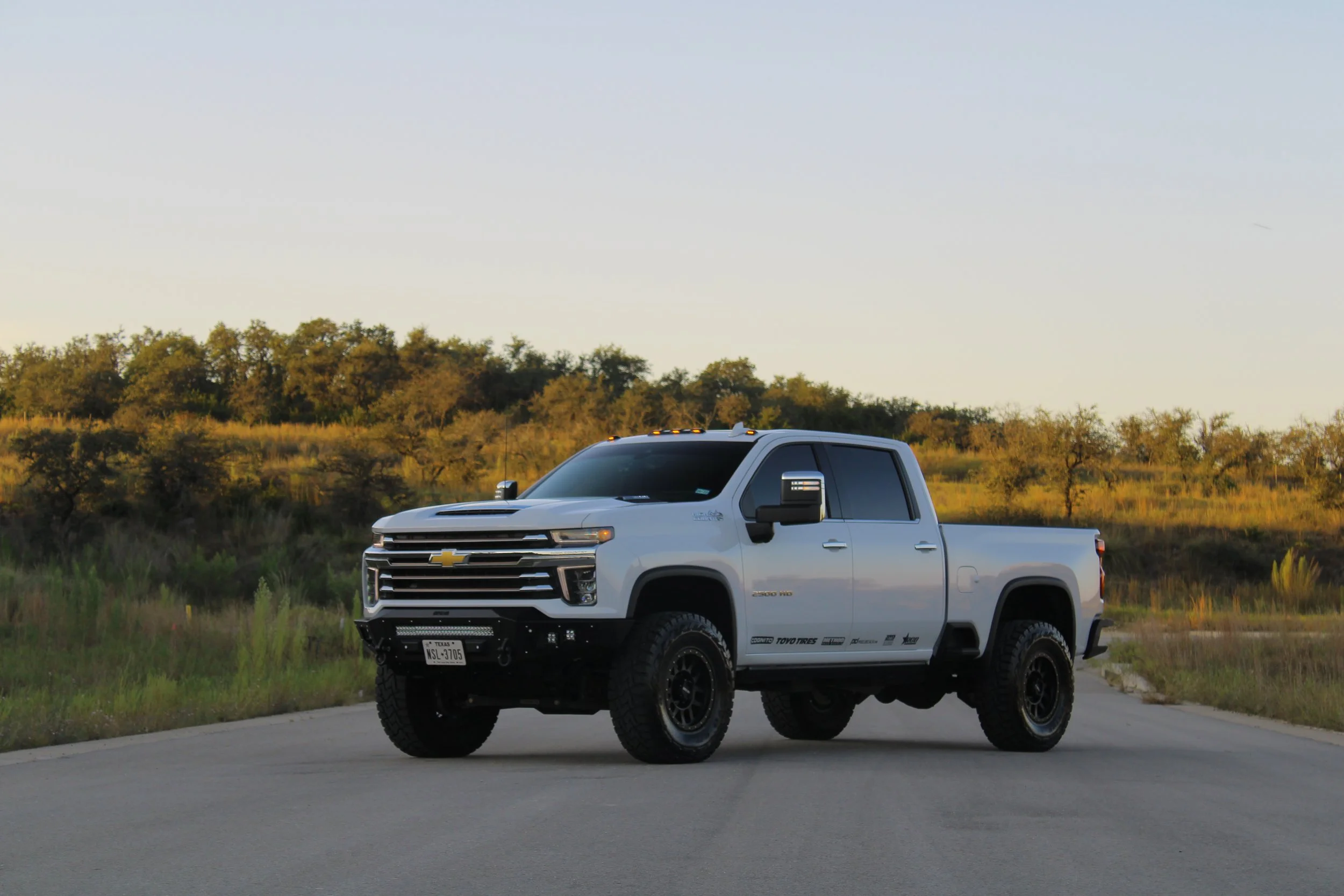 A white Chevrolet Silverado pickup truck parked on a paved road in a rural area during sunset, with green vegetation and trees in the background.