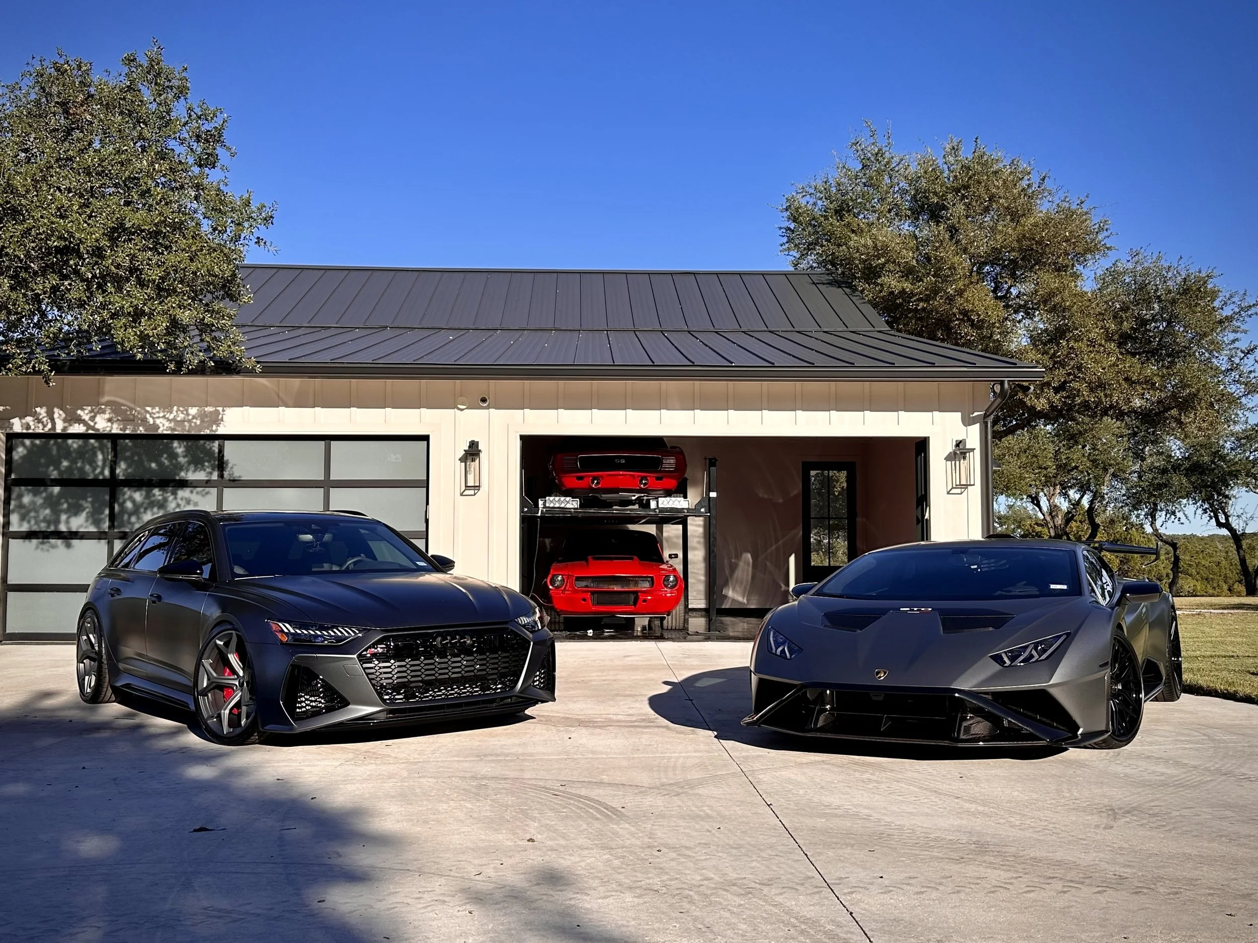 A driveway with two black luxury cars, a sedan and a sports car, parked outside a modern house with a garage. The garage has two shelves with red sports cars, and the house has a metal roof and large glass windows. Trees and a blue sky are in the background.