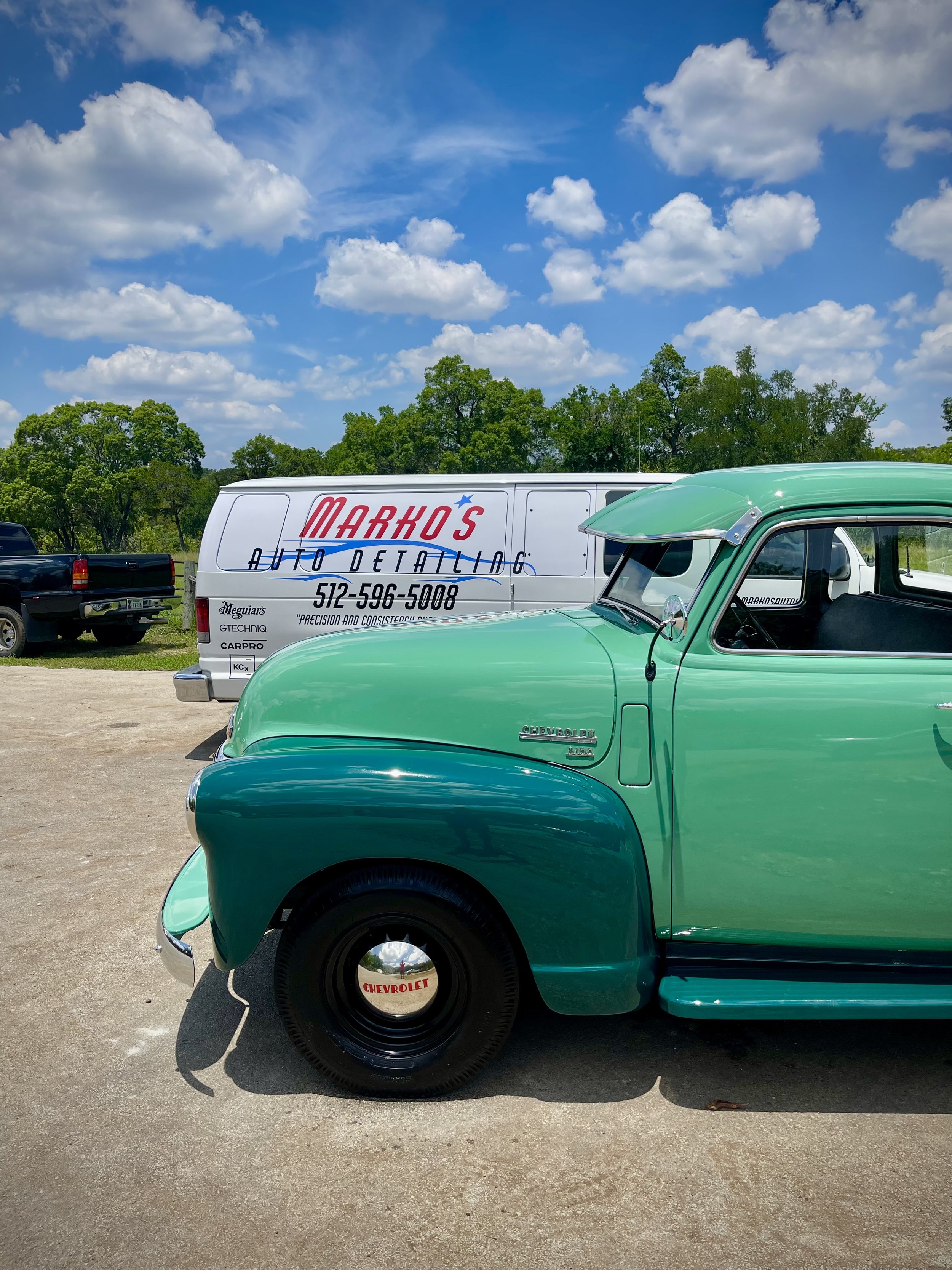 Vintage green Chevrolet truck parked on a gravel lot with a white auto detailing van and a black pickup truck in the background, under a partly cloudy blue sky.
