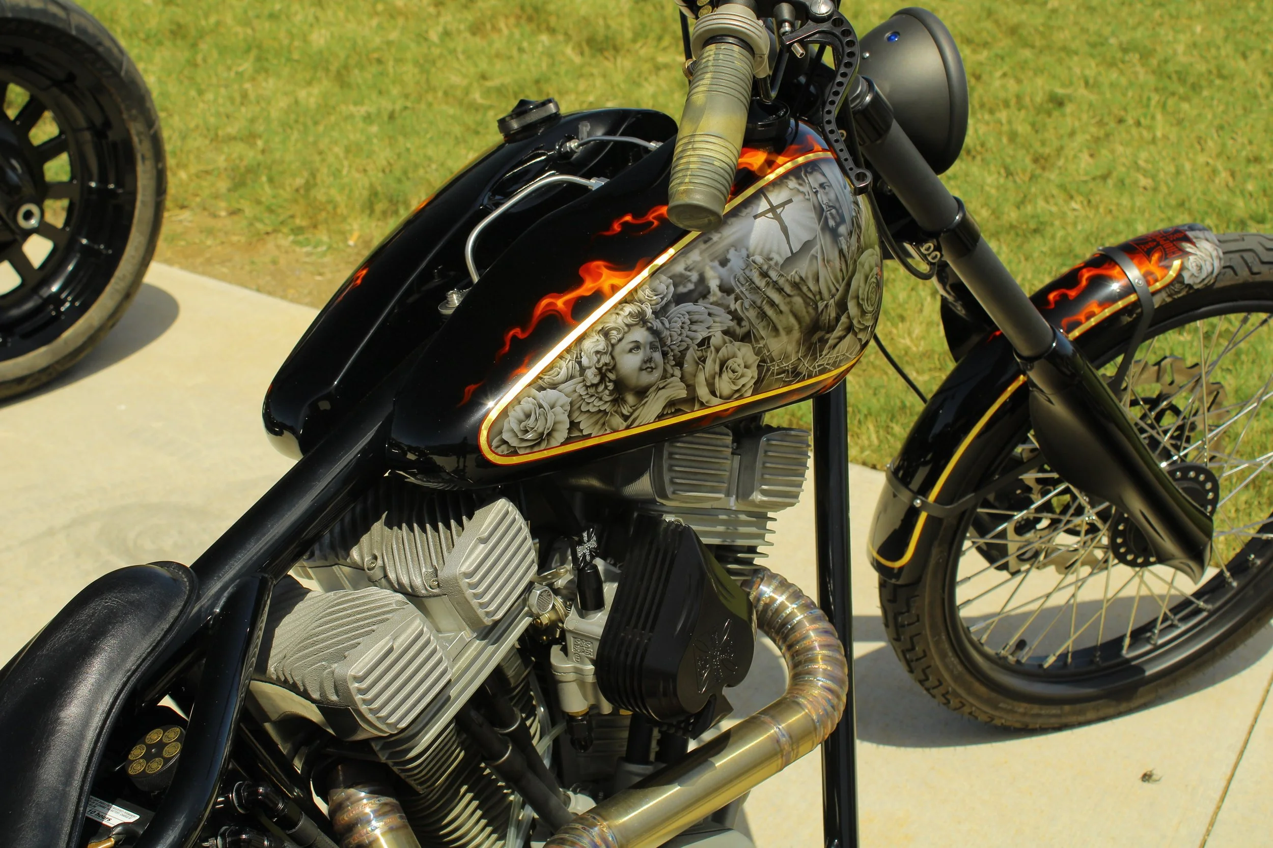 Close-up of a custom black motorcycle with detailed airbrushed artwork on the fuel tank, featuring religious imagery, angels, roses, and flames. The motorcycle has a V-twin engine, black frame, and spoked wheels, with a background of grass and concre