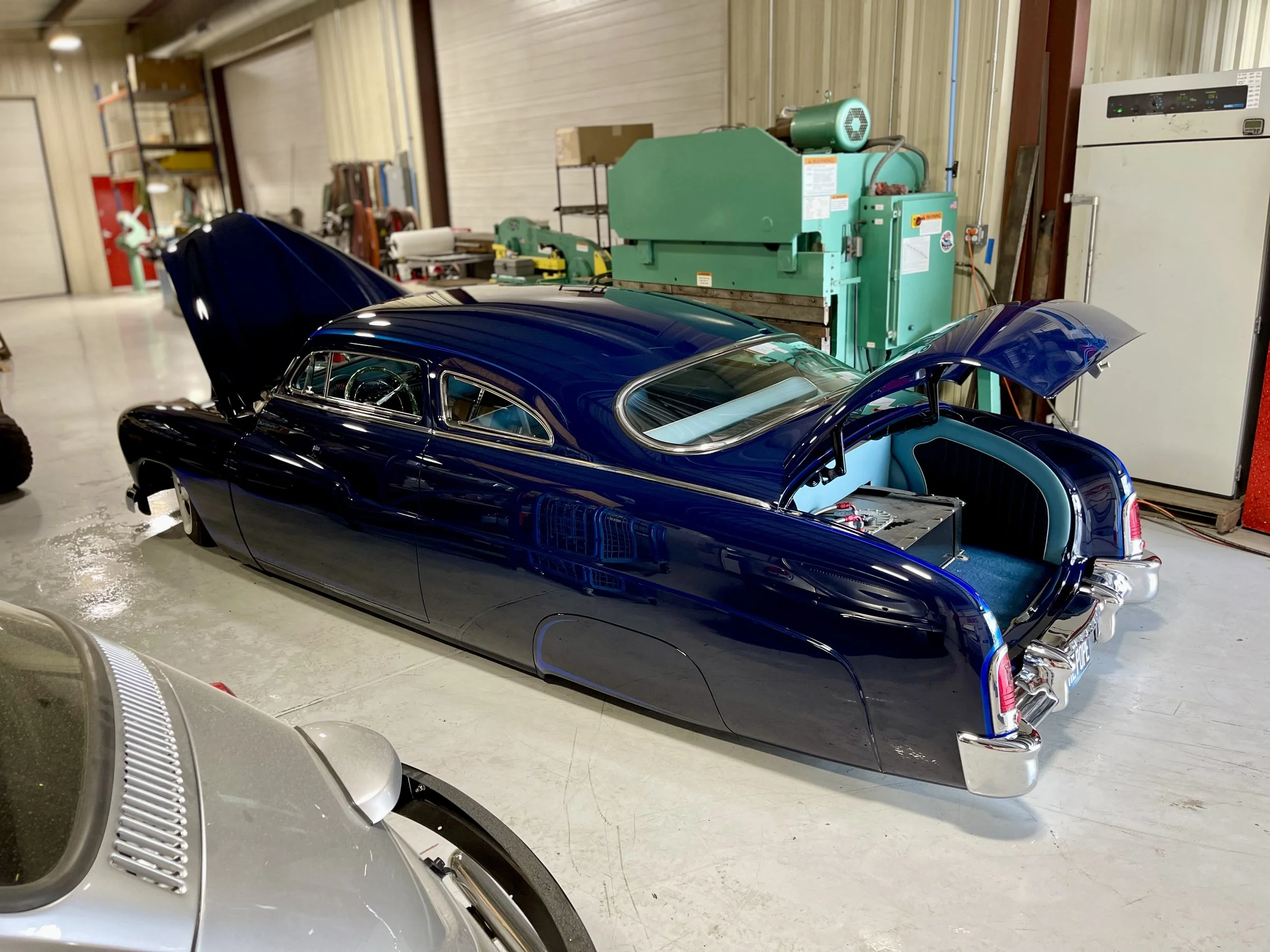 A vintage, dark blue car with its hood and trunk open located in a workshop or garage.