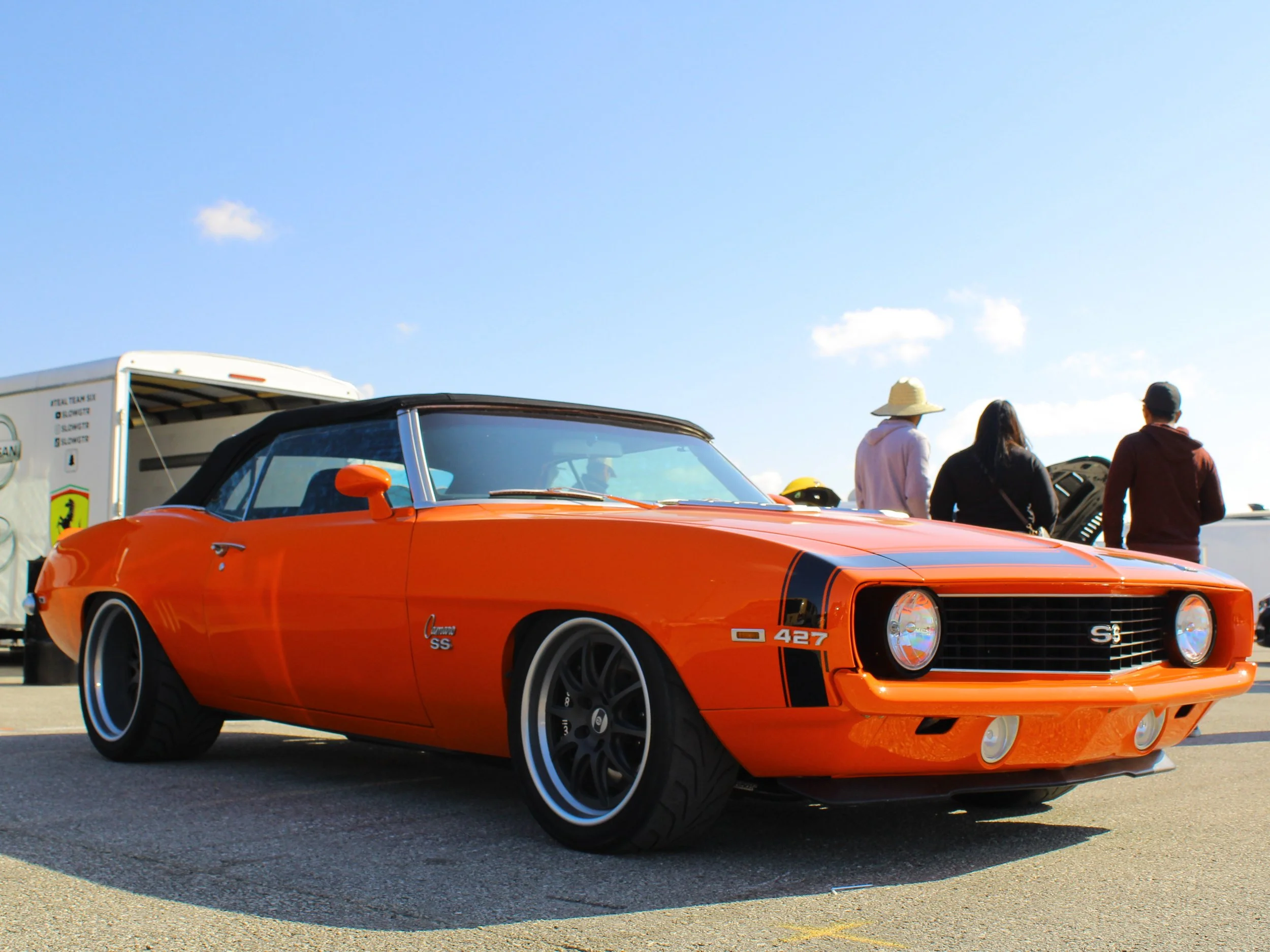An orange vintage Chevrolet Camaro SS with a black top parked outdoors against a blue sky, with a few people and a trailer in the background.