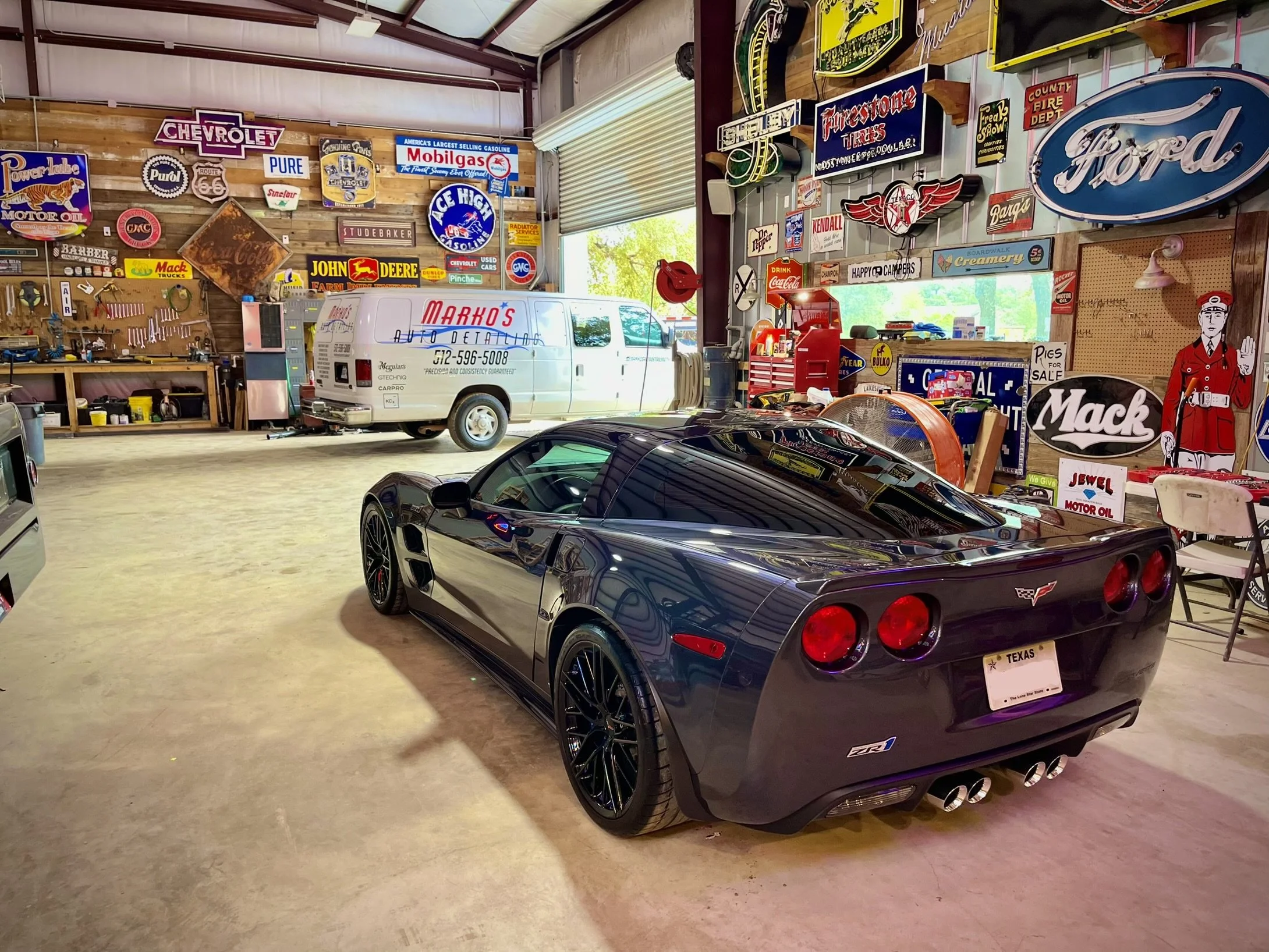 Black Chevrolet Corvette sports car parked inside a vintage garage filled with colorful retro signs, tools, and memorabilia.