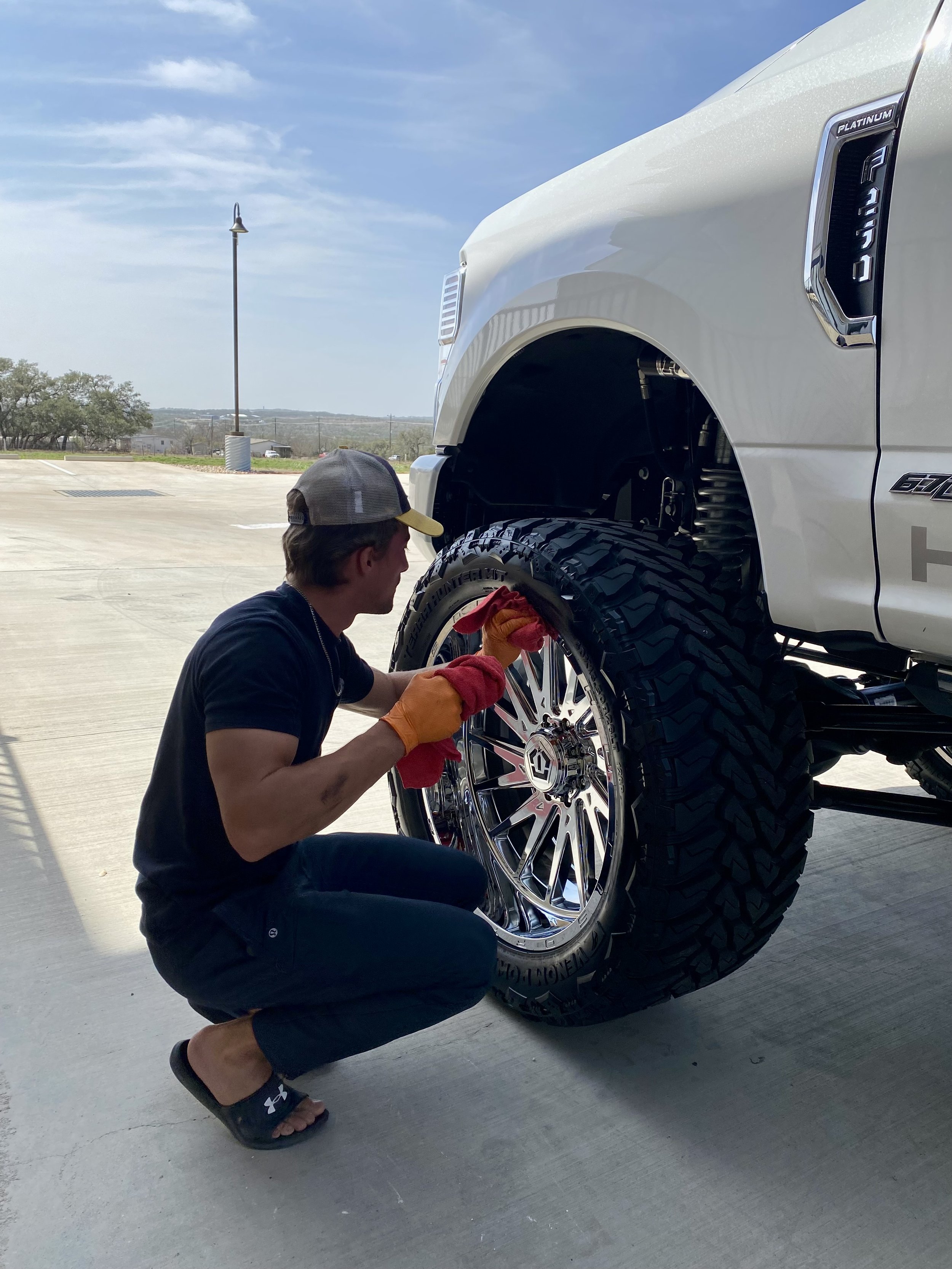 A man wearing a cap, black t-shirt, shorts, and orange gloves, crouching down, cleaning a large chrome wheel of a white pickup truck with oversized tires, in an open parking lot under a blue sky.