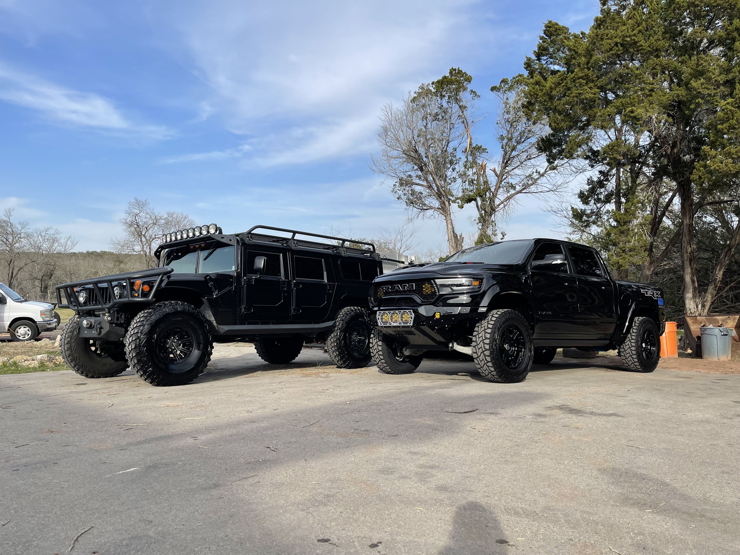 Two black trucks parked side by side on a paved lot, with trees and a blue sky in the background.