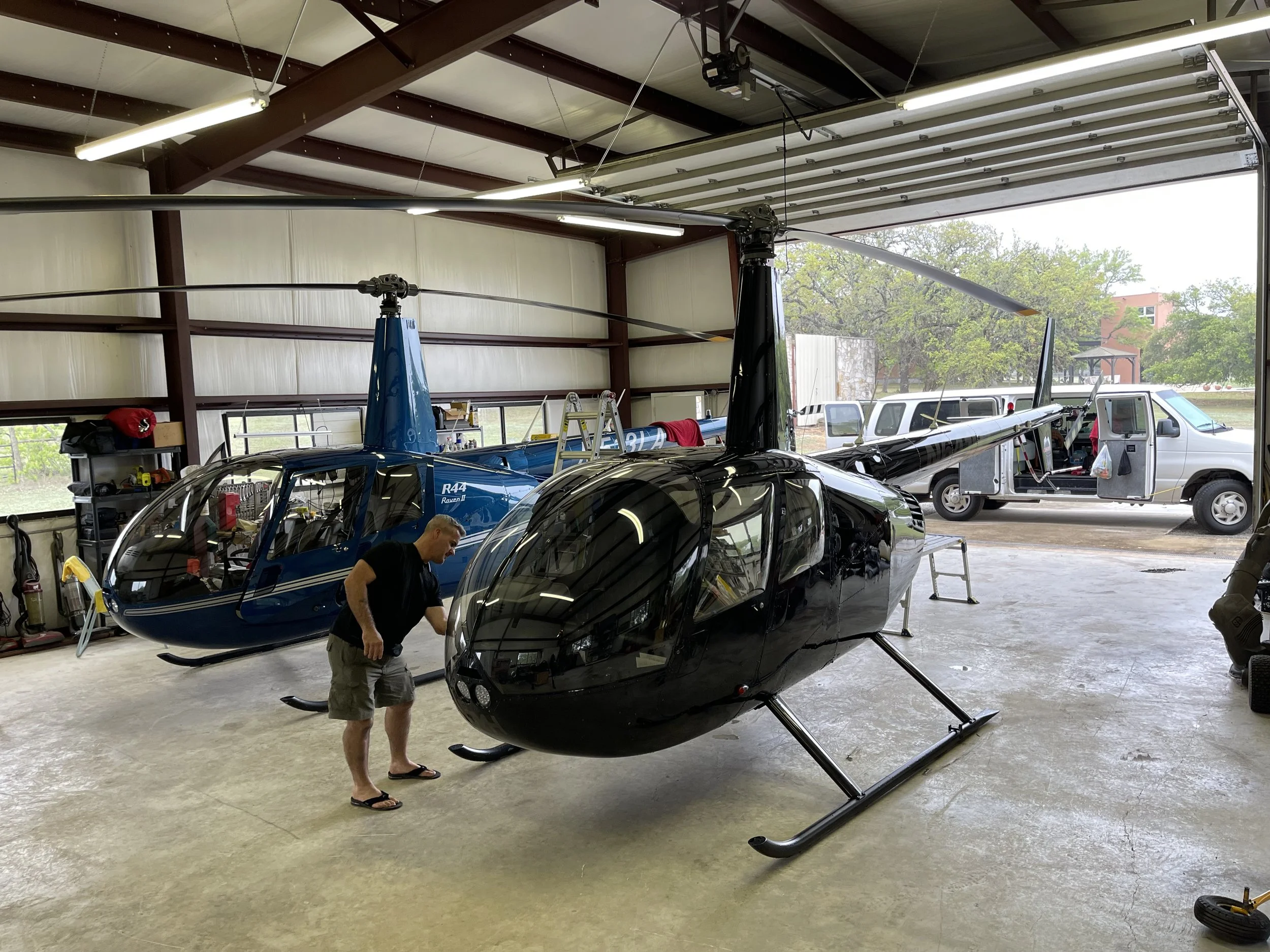 A man working inside a hangar with two helicopters. The black helicopter in the foreground and a blue helicopter behind it, with a service van outside the hangar visible through the open garage door.