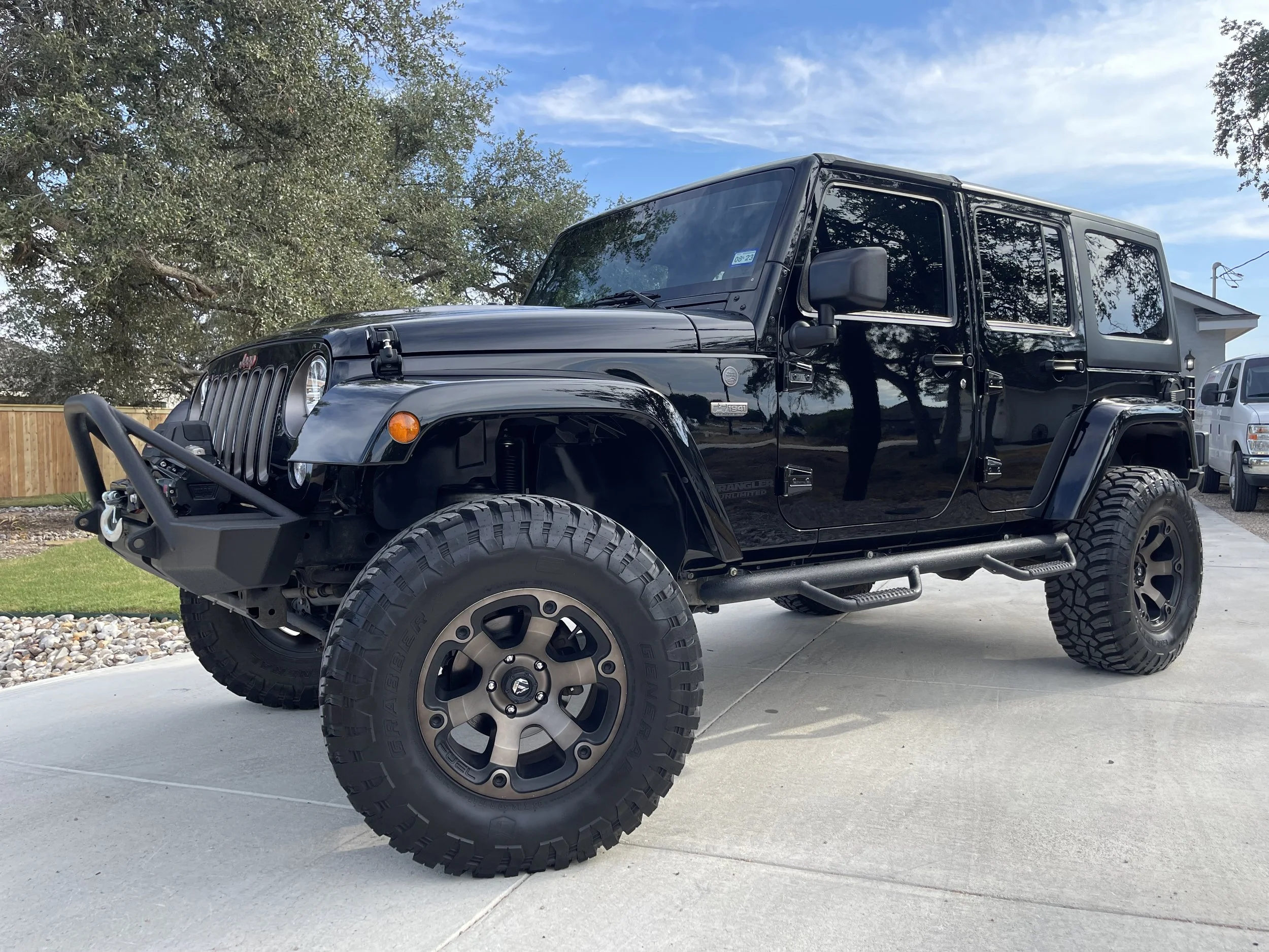Black Jeep Wrangler four-door SUV parked on a driveway with a wooden fence, trees, and a house in the background.