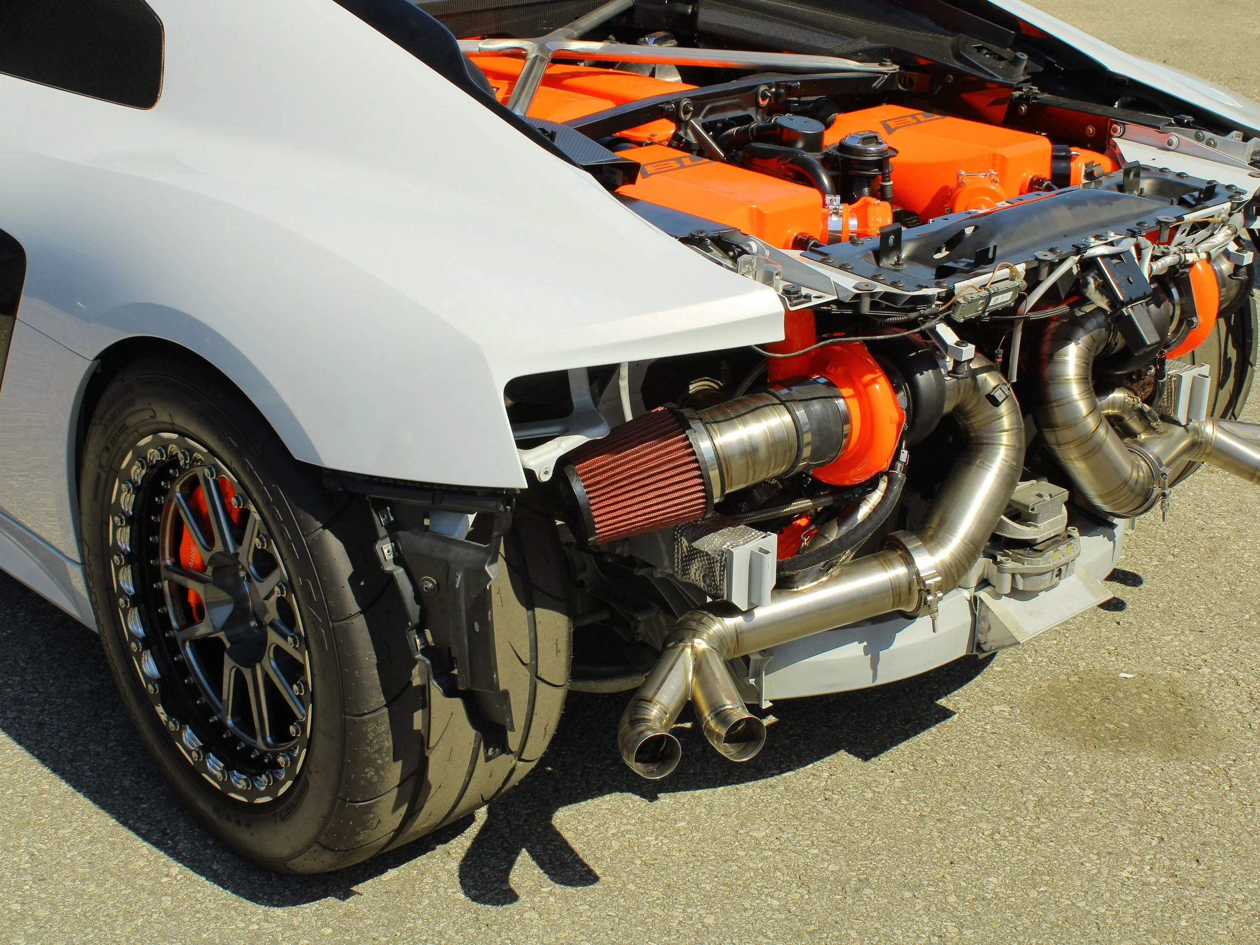 Close-up of a high-performance race car's front with exposed engine, racing slick tire, and orange accents.
