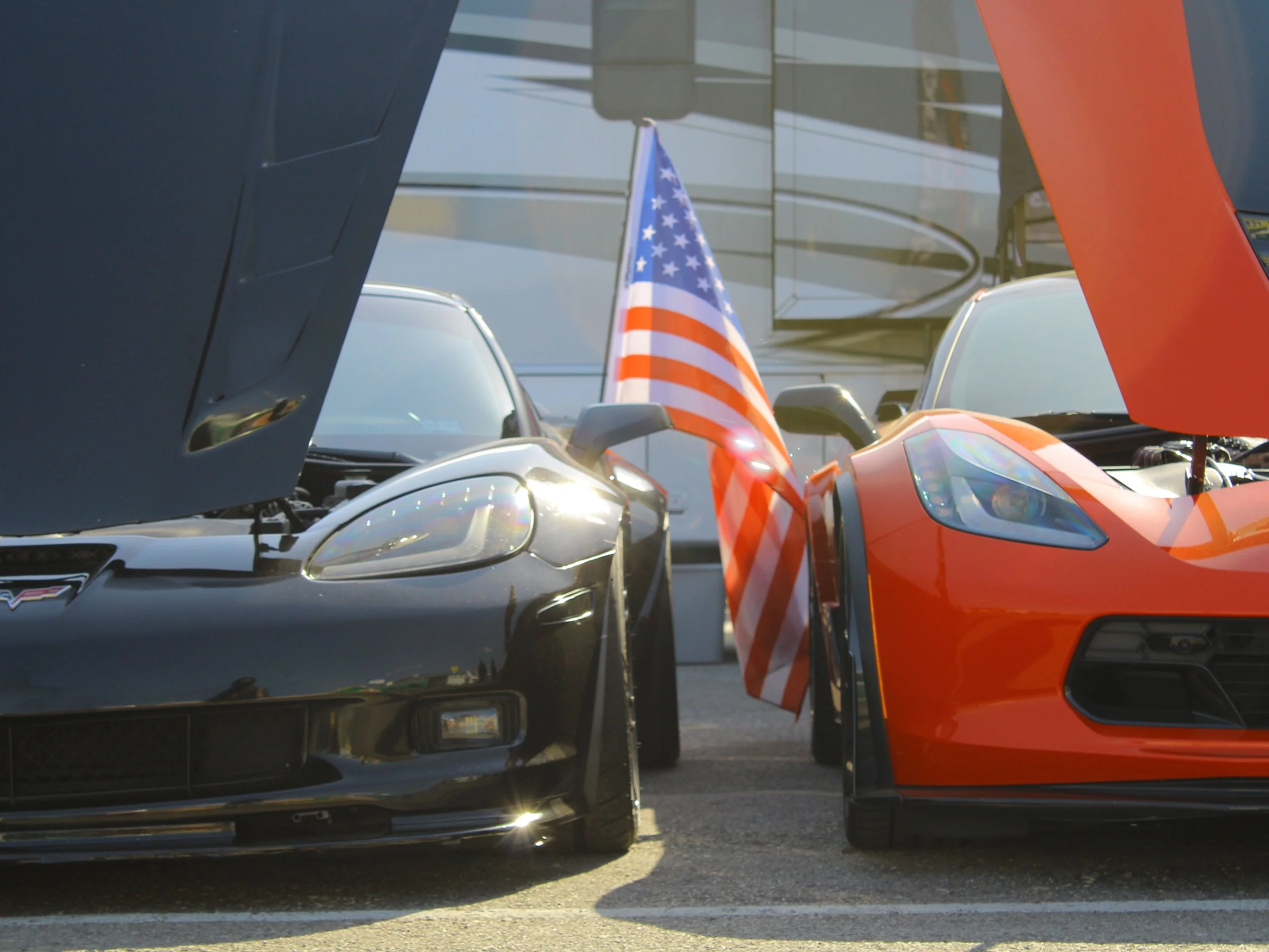 Close-up of two sports cars with open hoods, one black and one orange, parked side by side, with an American flag in between.