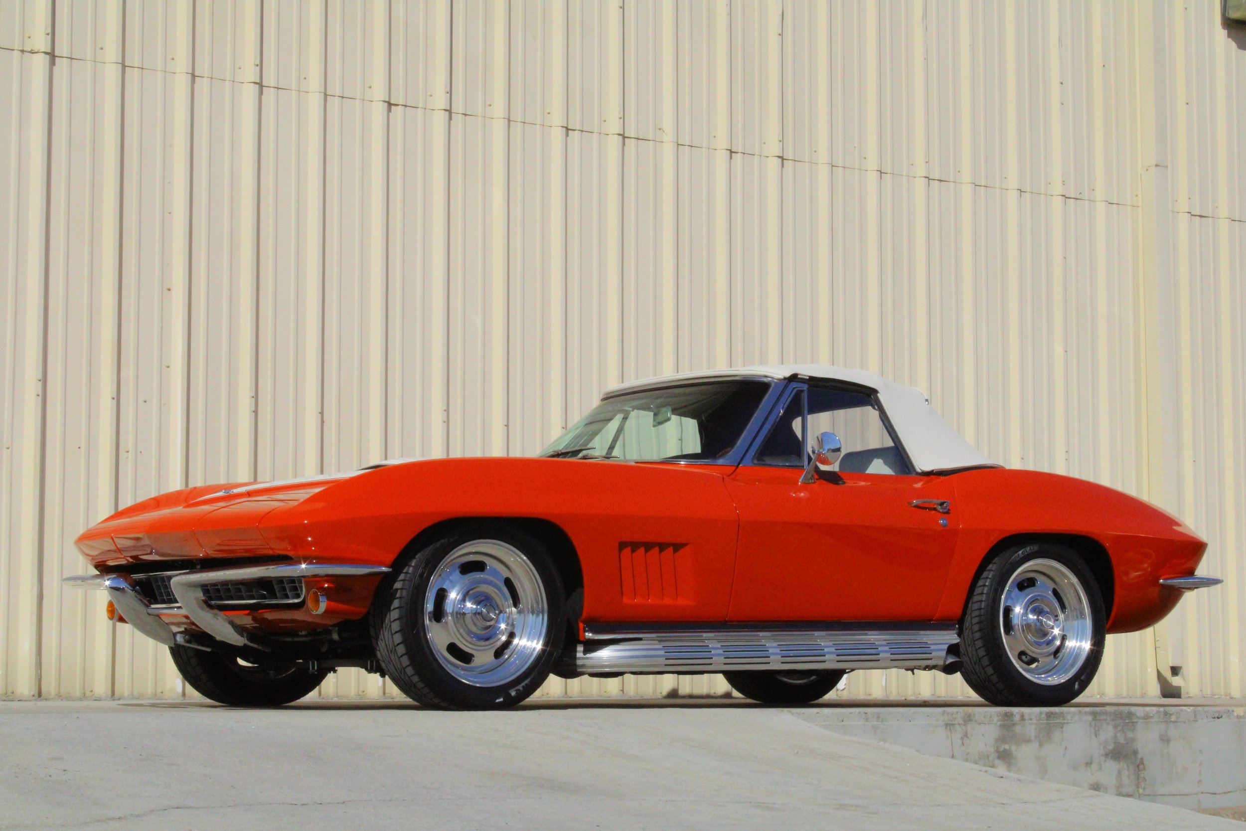 A red classic Chevrolet Corvette convertible with a white soft top, parked on a concrete surface in front of a beige metal building.