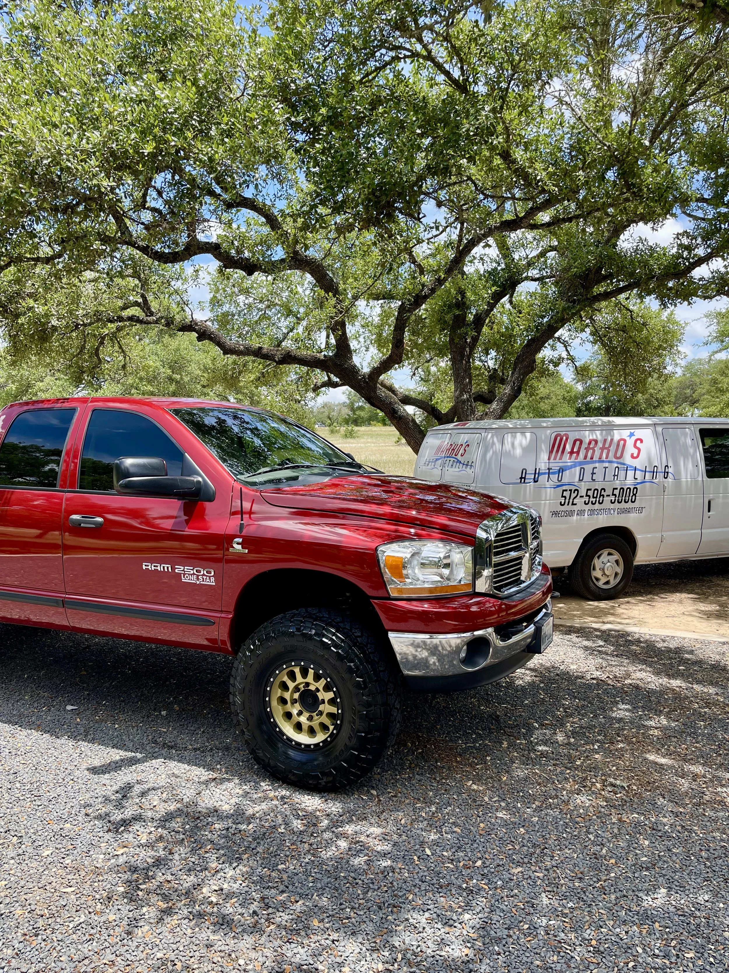 A red RAM 2500 Lone Star truck parked under a large green tree, next to a white Markos auto detailing van, on a gravel surface in an outdoor setting.
